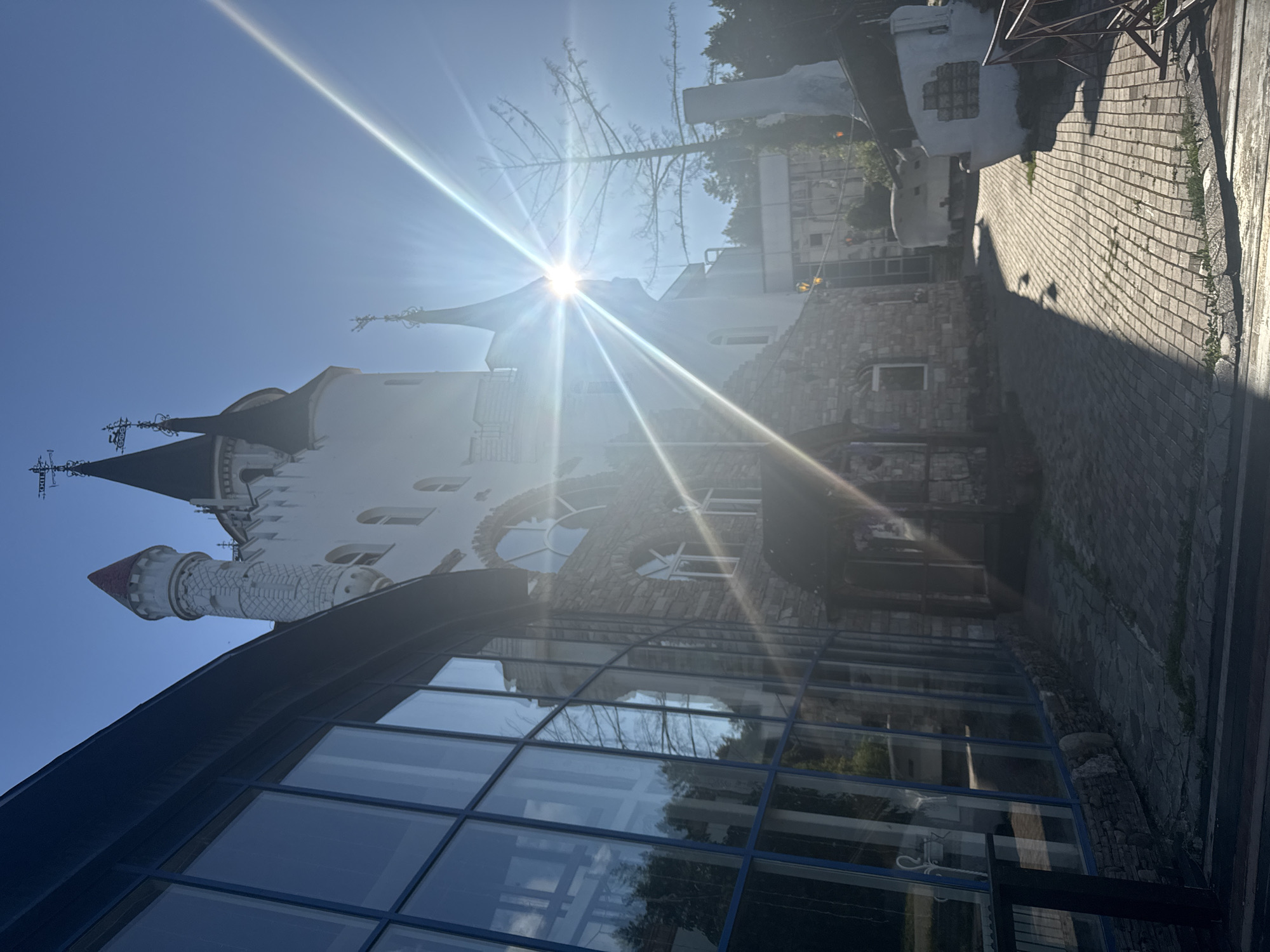 The image depicts the exterior of a whimsical, castle-like building with several towers, possibly part of a themed park or entertainment venue. The architecture features white walls, pointed roofs, and decorative elements. A large, modern glass-paneled structure is in the foreground on the left, reflecting the sky and surrounding trees. The ground is paved with cobblestones, and there are shadows cast by the sun. The sun is very bright, causing lens flare and obscuring details in the upper right quadrant of the image. The building appears to be situated in a natural, possibly wooded, environment. The location is identified as Ufa, Russia. There are no people visible in the image.