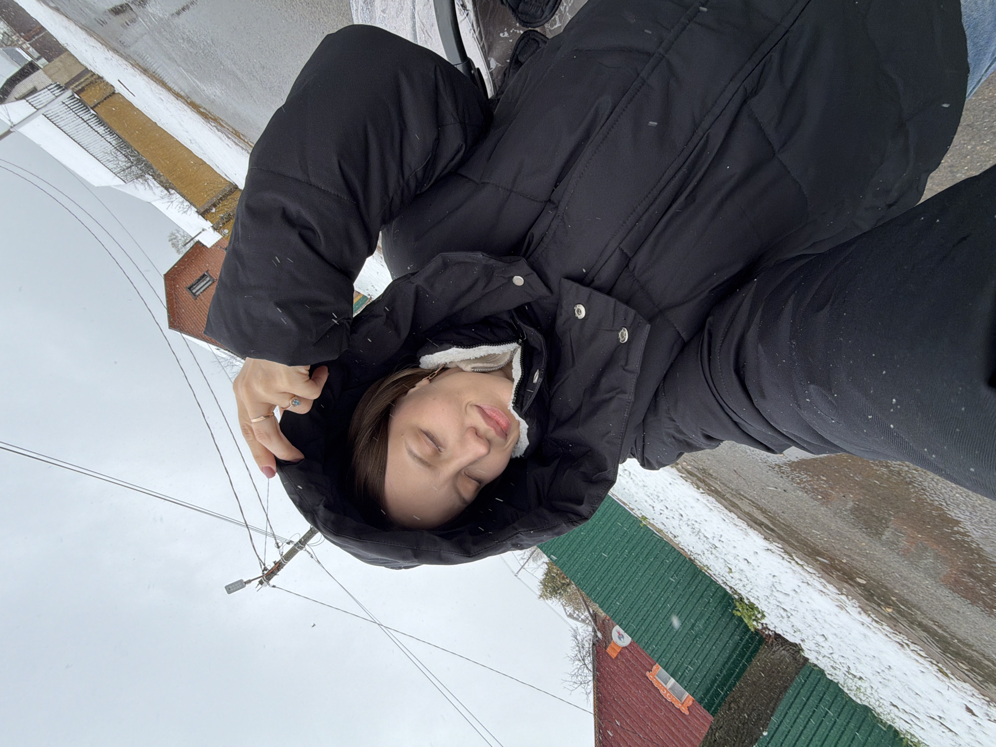 A young woman is depicted outdoors in what appears to be a winter setting, likely in Reshetnikovo, Russia. She is shown from the chest up, wearing a black, puffy winter jacket with the hood pulled up. A white, fluffy lining is visible around the collar of her jacket. Her eyes are closed, and her expression appears serene or peaceful, with her head slightly tilted. Her left hand, adorned with a ring, is positioned near her face, possibly holding onto the hood.
Light snow is falling, with small white flakes visible on her jacket, in her hair, and scattered throughout the air. The background features a residential or semi-rural environment. Patches of snow are visible on the ground, which appears to be a street or paved area. To the left of the woman, there's a building with a reddish-brown roof and siding, while to her right, another building has a distinct green metal roof. Overhead, power lines or telephone wires crisscross the overcast sky. The overall atmosphere suggests a quiet, snowy daytime scene. No visible text is present in the image.