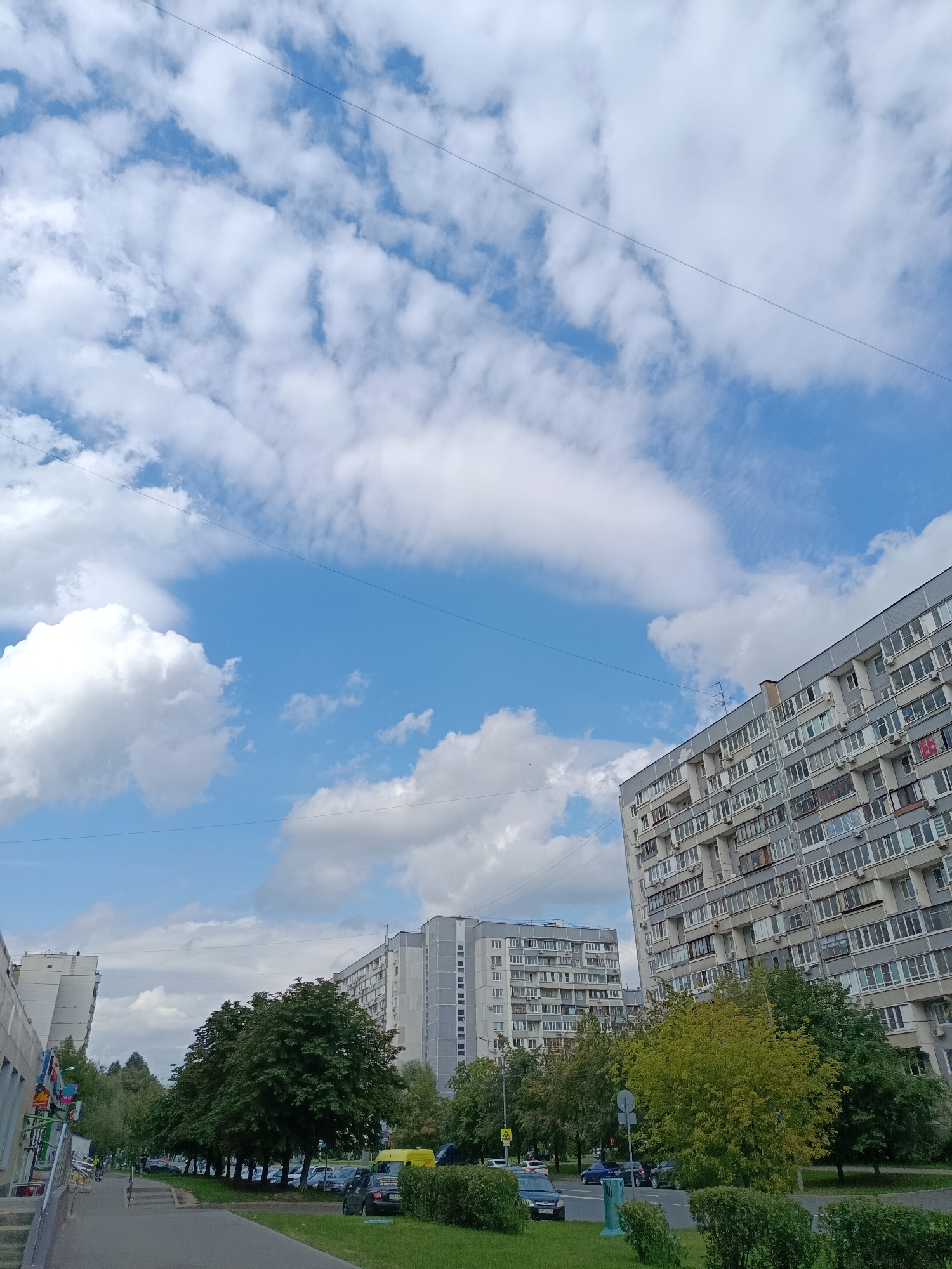 The image depicts a residential area in Moscow, Russia, under a partly cloudy sky. In the foreground, a sidewalk runs alongside a road with parked cars and lush green trees. Several large, multi-story apartment buildings, typical of Soviet-era architecture, dominate the background. The buildings are characterized by their uniform window layouts and balconies. One building, in the right foreground, is particularly prominent, its facade angled towards the viewer. Further down the street, more apartment blocks and trees are visible. A yellow van is parked near the curb, and a few individuals are seen on bicycles in the distance, indicating normal city life. The scene is bathed in daylight, suggesting it was taken during the daytime. The sky is a vibrant blue with scattered white cumulus clouds.