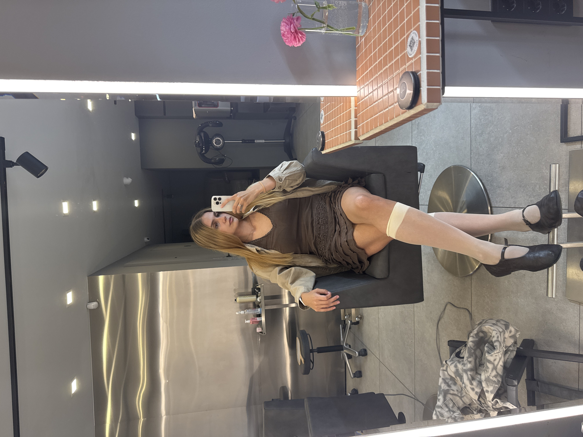 A young woman sits in a black chair in a hair salon, as reflected in a mirror. She holds a smartphone in her right hand, presumably taking a selfie. She wears a brown dress, a beige jacket, white stockings, and black shoes. The salon appears modern, with a stainless steel wall and various hair care products visible in the background. A tile-covered counter with a vase of pink flowers is positioned to the upper right. Based on the overall aesthetic and available information, it is likely that this photo was taken in Tbilisi, Georgia.
