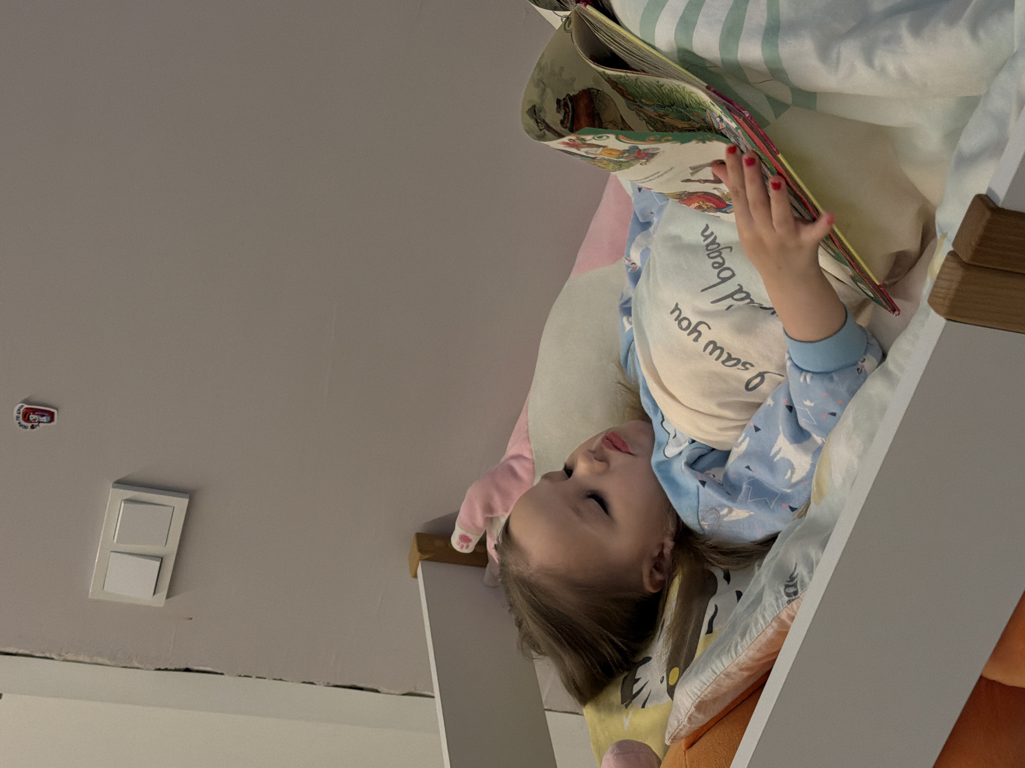 A young child is lying on their side in a bed, holding open a book. The child has fair skin and brown hair. They are wearing a blue and white patterned pajama top and have red nail polish on their fingers. The book is open to a page with illustrations. The bedding includes a variety of colors and patterns, including stripes, animal prints, and solid colors. The bed appears to be a bunk bed or a high bed, with a white side rail visible. The background shows a light gray wall with a light switch and a small sticker. The location is not specified as the city of Chaikovsky, Russia, in the image.