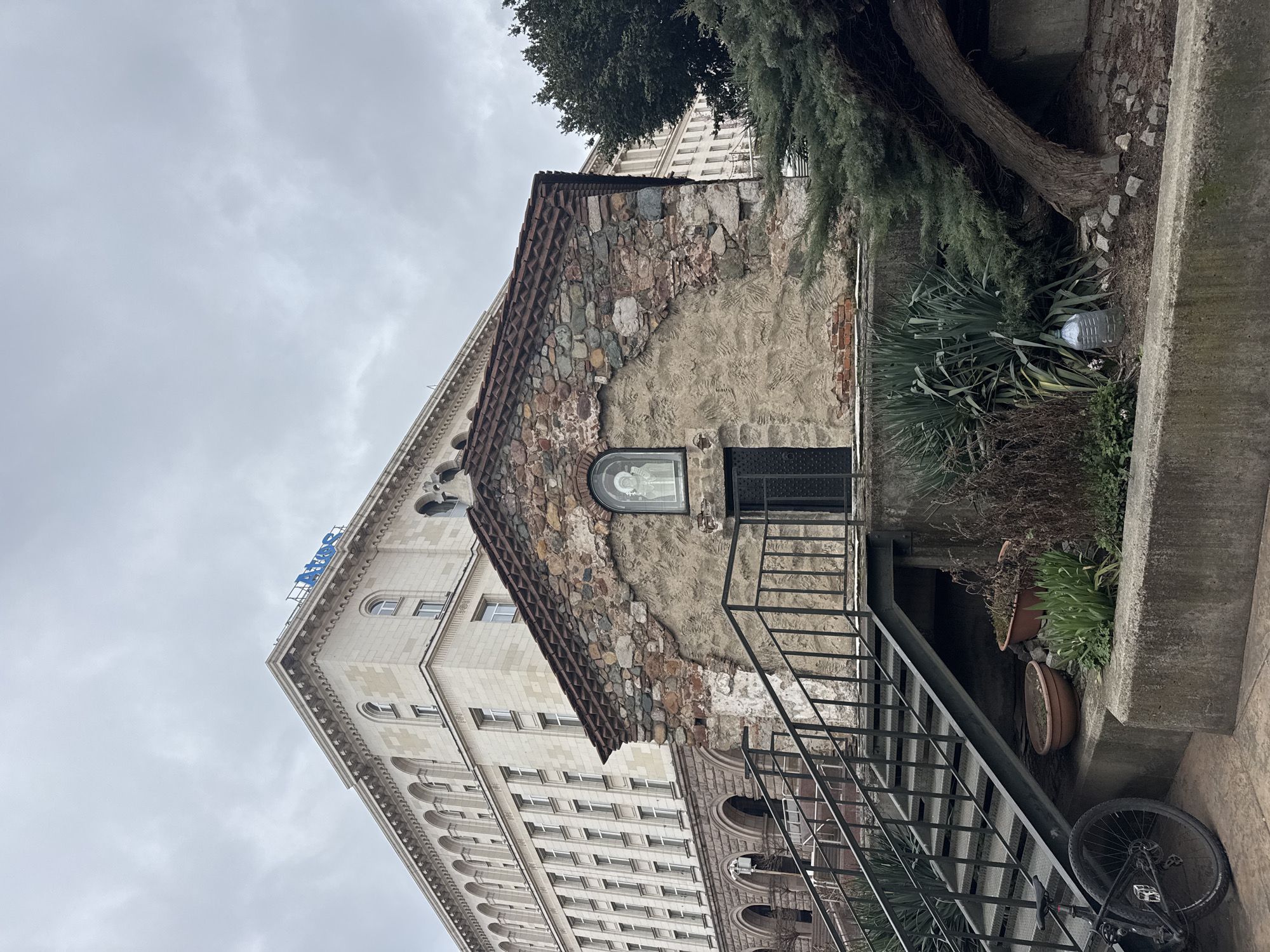 The image captures a view of the Church of St. George in Sofia, Bulgaria, an early Christian red brick rotunda. The building adjoins a larger, multi-story modern building with a sign that says "ЦУМ" (TSUM). The roof of the church is visible, partly constructed of stone and partly covered in reddish-brown tiles. The facade is peeling away, revealing the underlying stone construction. A small window with a religious icon can be seen. A black metal staircase leads to a lower level. To the right of the church, there's greenery and a tree. A bicycle leans against a concrete wall near the bottom right of the frame. The sky is overcast.