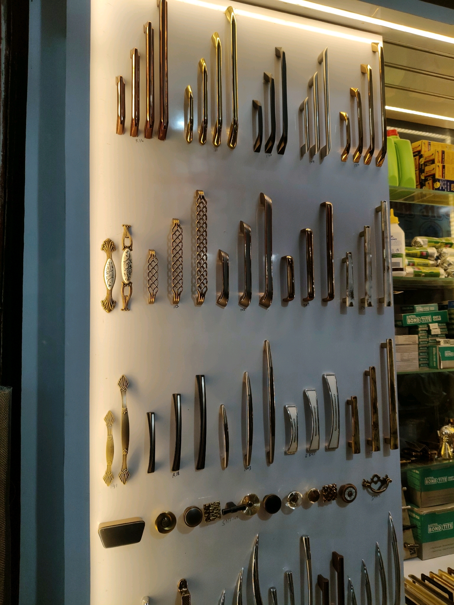 The image depicts a retail display of various door and cabinet handles in a store located in Darbhanga, India. The handles are arranged in neat rows on a white board, showcasing a range of styles, materials, and finishes, including polished brass, copper, and brushed metals. Some handles are simple and modern, while others feature ornate designs and decorative elements. The display is illuminated by overhead lighting, highlighting the metallic sheen of the products. In the background, shelves stocked with packaged goods, including bottles and boxes, are visible, suggesting a hardware or home improvement store. The scene is static, with no people or ongoing actions evident, focusing solely on the presentation of the merchandise.