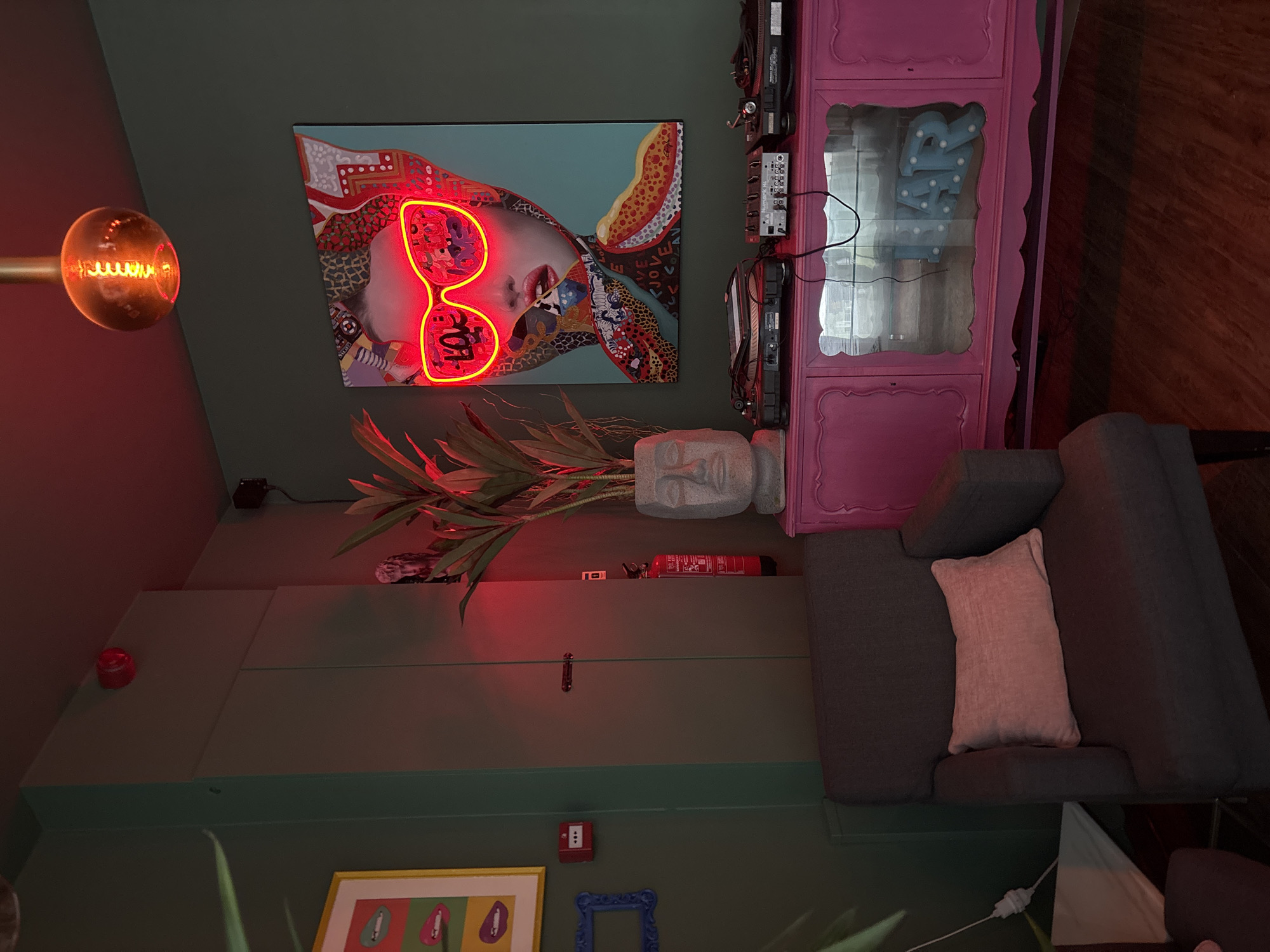 The image shows an interior space in what is possibly a room in Lisbon, Portugal, given the style of decoration and the types of objects present. The room features a gray sofa with a light-colored pillow, a pink cabinet, a painting on the wall depicting a woman with neon-light sunglasses, a lamp with an exposed filament bulb, a stone bust, a fire extinguisher, and other decorative elements. The walls are painted in green shades. The image suggests a modern, eclectic style, mixing art, design, and practical elements.