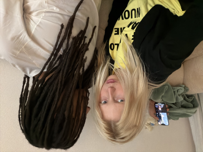 An indoor scene in Paris, France, depicts two individuals seated on a ribbed, light-colored sofa against a plain light wall. On the left, a fair-skinned individual with long blonde hair, wearing a black top over a yellow shirt with partial text (including "LOVE," "ME," "AI," and "NUOVO"), looks directly at the camera with a stern expression. This individual holds a black smartphone in their right hand, displaying an active video call or video content with at least two smaller video feeds visible on screen. Behind them, a crumpled green textile rests on the sofa. On the right, an individual with long, dark dreadlocks is seated, turned away from the camera, and wears a plain, light-colored (likely white) garment.