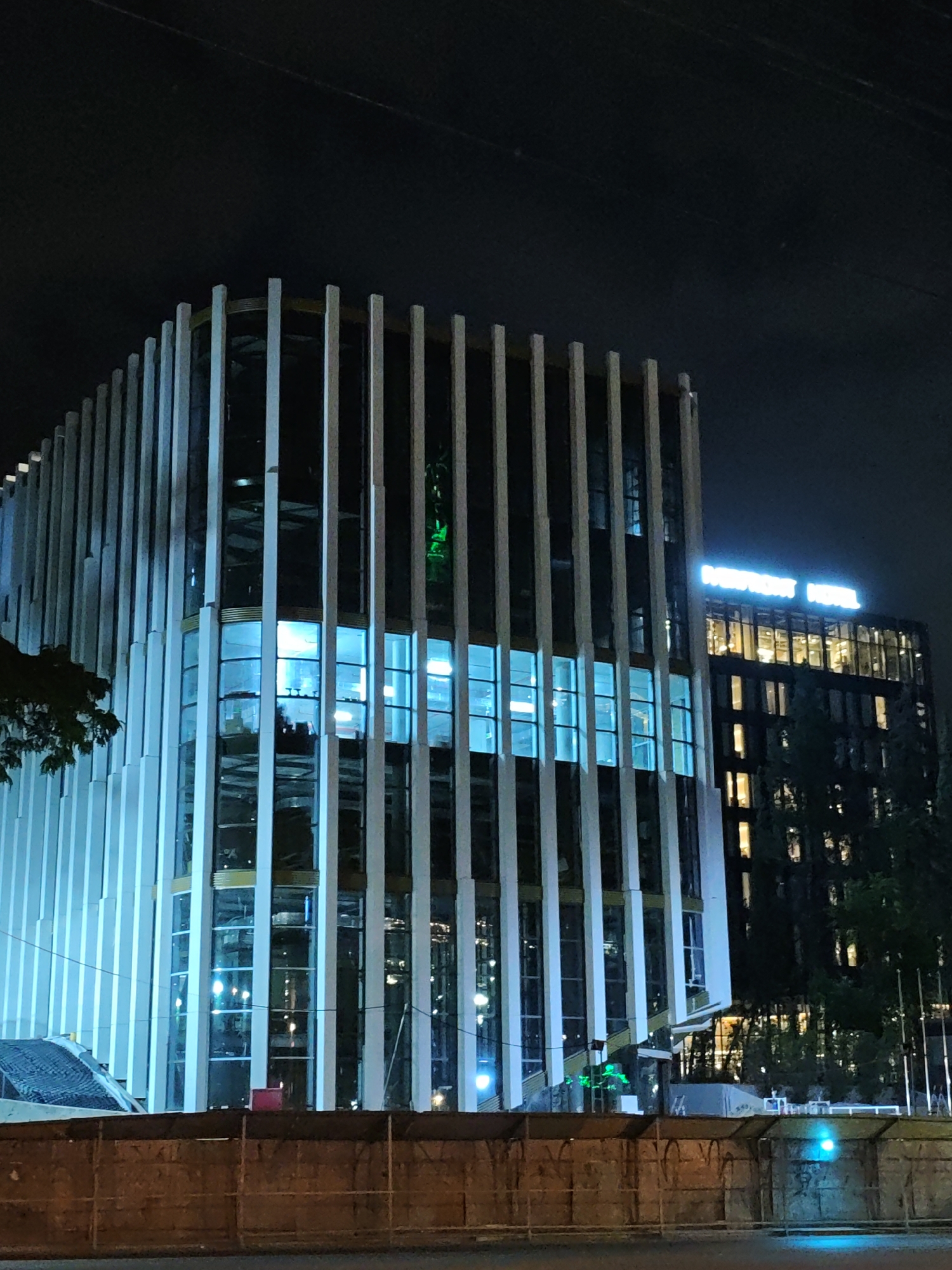 The image, taken at night, shows two modern buildings in Tbilisi, Georgia. The foreground is dominated by a dark, rustic-looking fence, with some blue light reflecting off it. Behind the fence, the first building is a sleek, multi-story structure with a facade of vertical white pillars and large glass windows. The windows are illuminated with a bright, cool blue light, giving the building an ethereal glow. Some of the interior can be seen through the glass, with multiple floors and some furniture visible. A tree with dark foliage partially obscures the left side of this building.
To the right of the first building, a second, taller building stands. This building has a darker exterior and is illuminated by numerous windows, each glowing with a warm, yellowish light, suggesting occupancy. At the very top of this second building, a prominent neon sign glows in a bright blue hue, spelling out "NEPTUNE HOTEL". The sky above is dark and featureless, typical of a night scene in an urban environment. The overall impression is one of contemporary architecture set against the backdrop of a night sky.