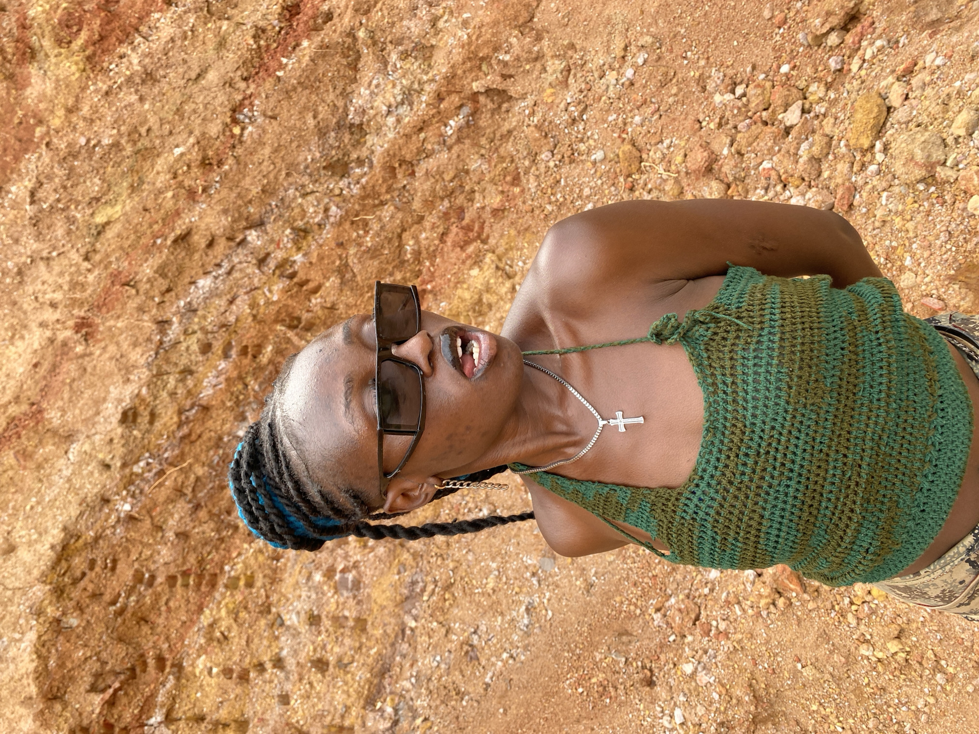 A young Black woman is captured from a low angle, tilted slightly to the right, posing against a backdrop of reddish-brown dirt and scattered rocks. She is wearing large, dark sunglasses and a green crochet top with varying shades of green. A silver chain with a cross pendant rests on her chest. Her hair is styled in braids, some of which have streaks of blue. Her mouth is slightly open, revealing her teeth and tongue. The scene appears to be outdoors during the day, with natural lighting and a dry, earthy environment. The location is identified as Bar Jirgi Summa, Nigeria.