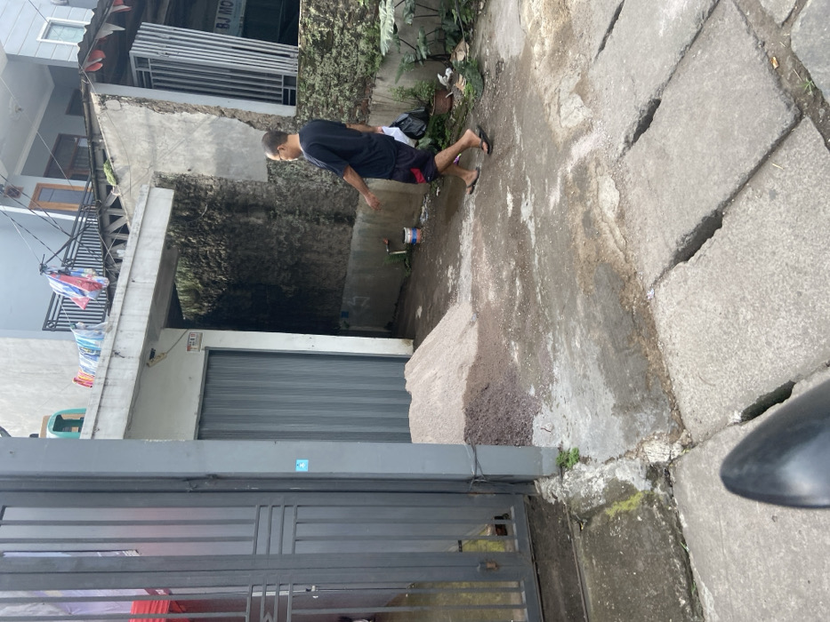 A man wearing a black t-shirt, red shorts, and black sandals walks to the left in front of a building. He is carrying a plastic bag. The building is a two-story residence, with a grey facade and a corrugated metal garage door. The image was taken in Bojongsoang, Indonesia, at the coordinates -6.979, 107.635. There is a pile of dirt on the ground in front of the garage door. There is a green plant growing in the dirt. The man is walking on a concrete surface. There are a series of drainage grates to the right of the man. There is a small blue container to the right of the garage door. The scene appears to be a residential neighborhood.