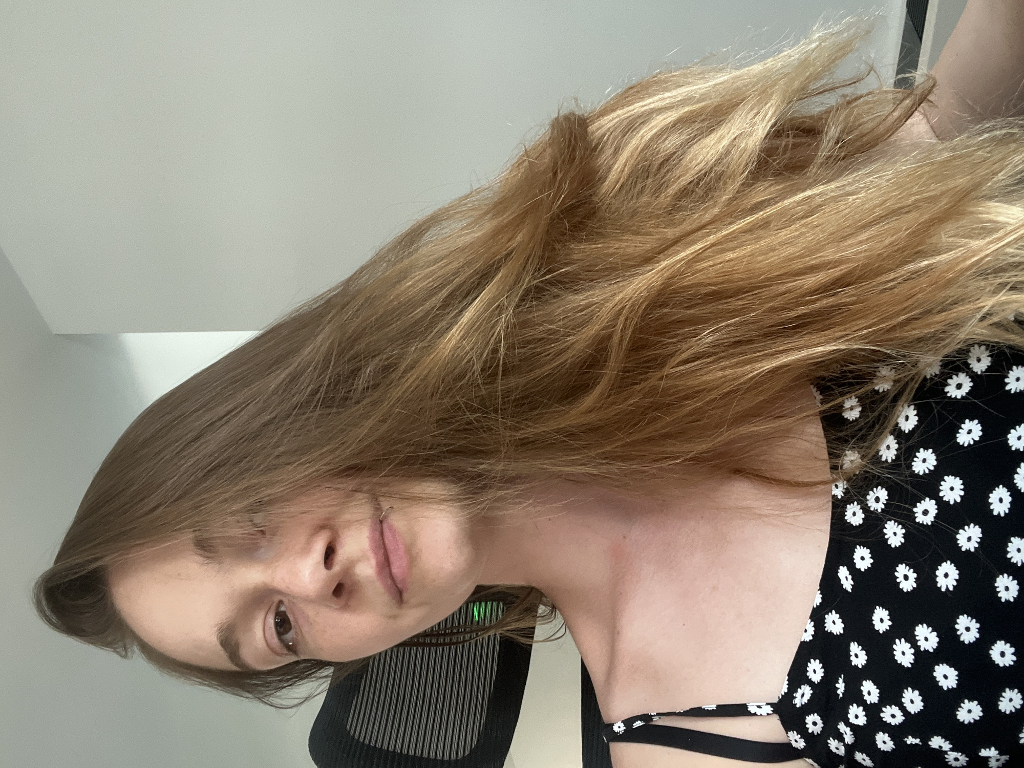 The image is a medium shot of a young woman with long, light brown hair, looking towards the camera. She is wearing a black dress with a white floral pattern and a black strap. Her face is slightly tilted, and her hair is falling over her right shoulder. The background is a plain, light-colored wall with a white corner visible on the left. A portion of a black office chair with horizontal lines is visible behind her left shoulder. A small, circular silver ring is visible on her lower lip. The scene appears to be indoors, possibly an office or a room. The specific location in the city of Plovdiv, Bulgaria, cannot be determined from the provided image.