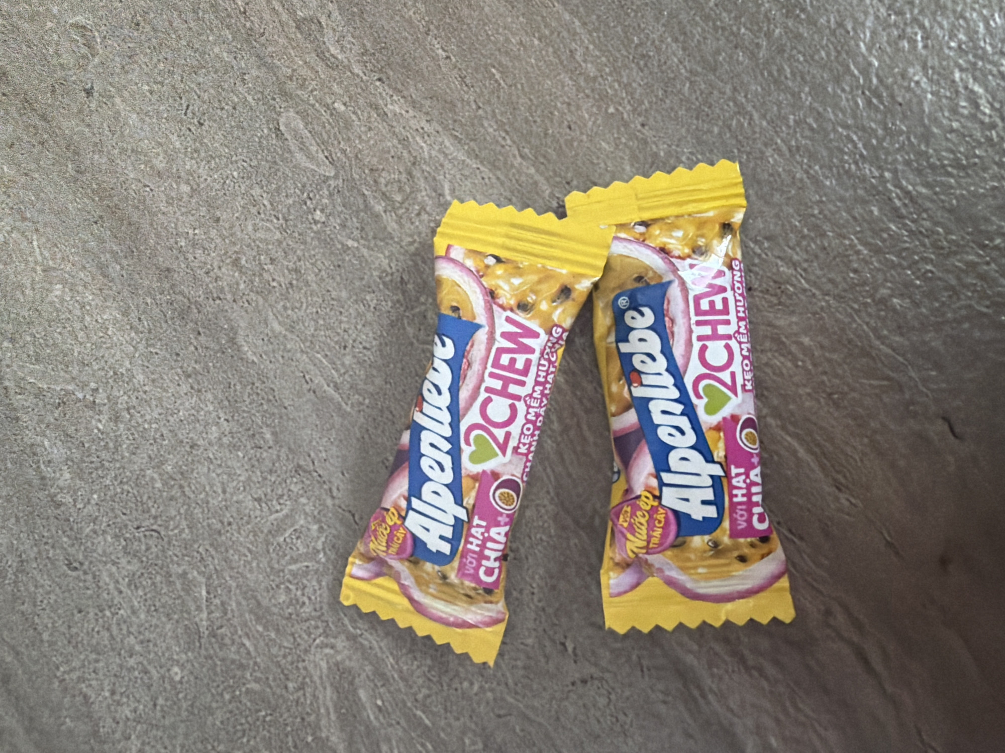 Two Alpenliebe 2Chew candy wrappers, each featuring a bright yellow background with graphics of passion fruit slices, are lying on a textured, grayish-brown surface. The wrappers are identical, with the Alpenliebe logo prominently displayed in blue with white lettering, accompanied by the "2Chew" branding in pink and green. Text in Vietnamese, including "Với hạt chia+" (with chia seeds) and "Kẹo mềm hương chanh dây, hạt chia" (soft candy passion fruit flavor, chia seeds), is also visible. The candy wrappers are positioned at an angle, with the top one slightly above and to the left of the bottom one. The environment appears to be an indoor setting with a surface that resembles concrete or a similar textured material. There are no people or animals present in the image. The image is taken from a top-down perspective. No specific location in Yuzhno-Sakhalinsk, Russia can be identified from this image.