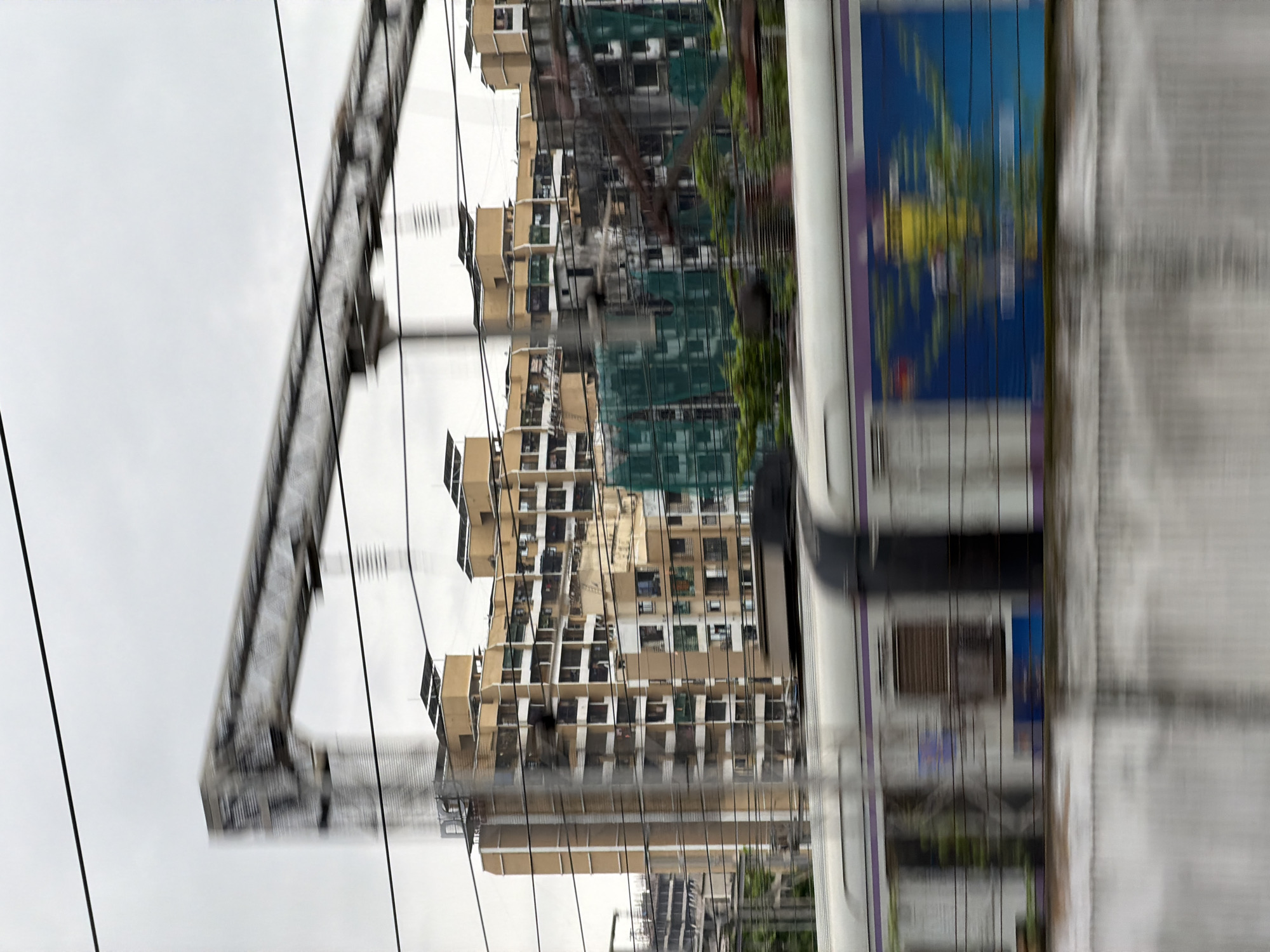 The image shows a blurred view of buildings and a train in Vasai-Virar, India. In the foreground, a train moves from left to right, displaying a colorful advertisement on its side. The train cars are predominantly white with purple trim. In the background, there are several apartment buildings, some of which appear to be under construction, covered with green netting. A large metallic structure, possibly part of an elevated railway line or overhead wires, is visible in the upper portion of the image, also blurred due to motion. Overhead power lines crisscross the frame. The sky is overcast, contributing to a muted color palette. The overall impression is one of urban transit and development.