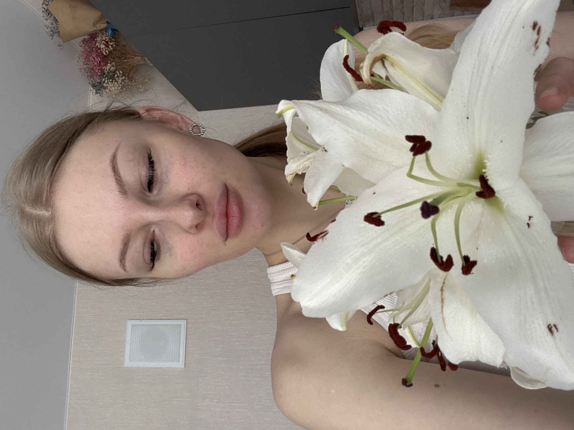 A woman is holding a bouquet of white lilies. She has fair skin, blonde hair, and an earring in her left ear. The woman is wearing a white top with a design on the shoulder. The background includes a wall with a small white air vent and a bouquet of dried flowers on a table in the background. It is difficult to determine the specific location in Perm, Russia from the provided image.