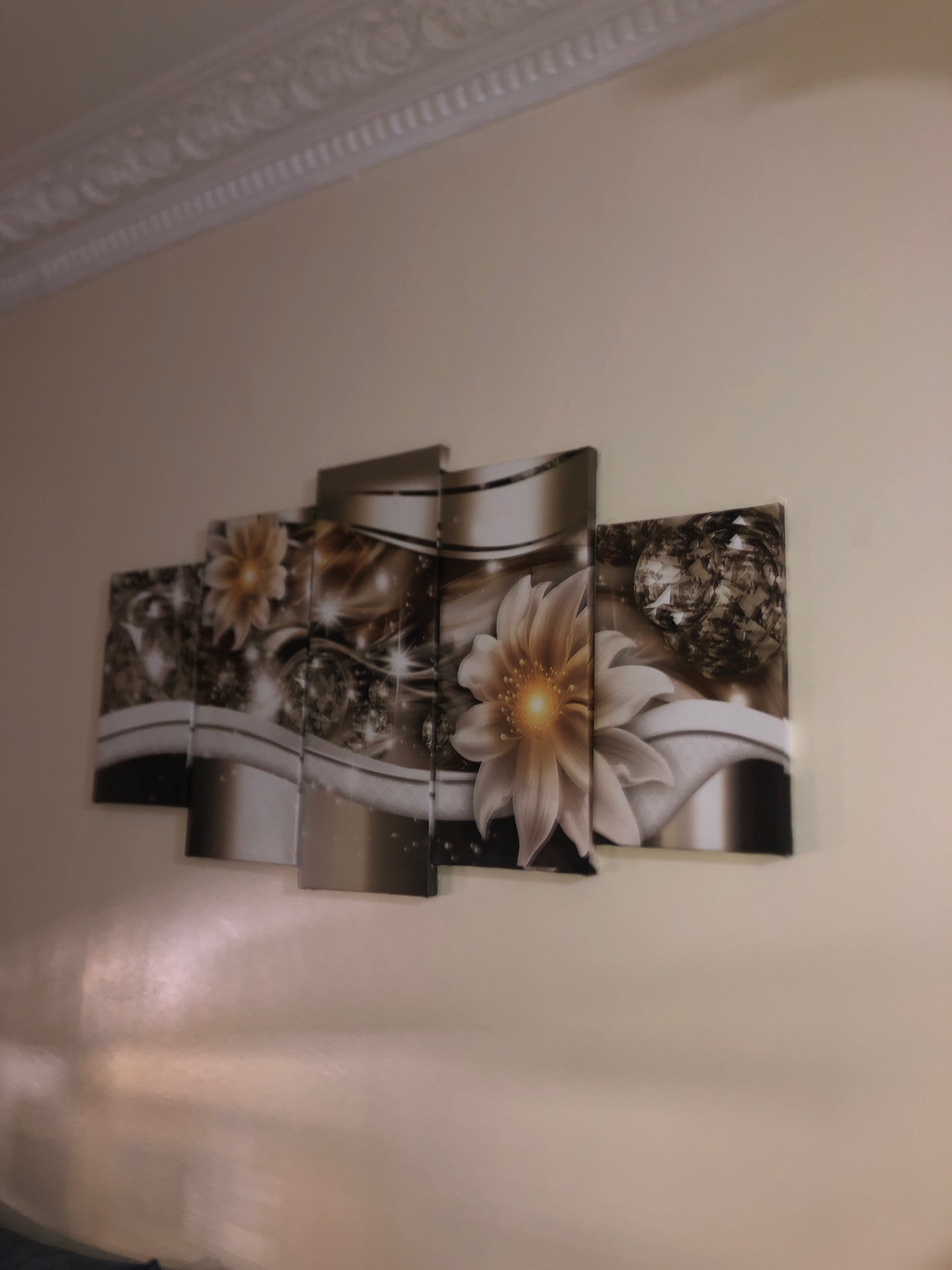 A five-panel abstract artwork depicting floral and crystalline elements is mounted on a light-colored wall. The artwork features a light-colored flower with a yellow center in the foreground, positioned on the second panel from the left. Behind the flower are swirling patterns and what appear to be large, faceted crystals, rendered in shades of brown, gold, and silver. The artwork is mounted at eye level in a domestic interior. Above the artwork, a decorative white cornice molding runs along the top of the wall. The lighting in the room creates a soft glow and reflections on the wall below the artwork. There are no people or animals visible in the image. The scene is consistent with a residential interior in Khemisset, Morocco.