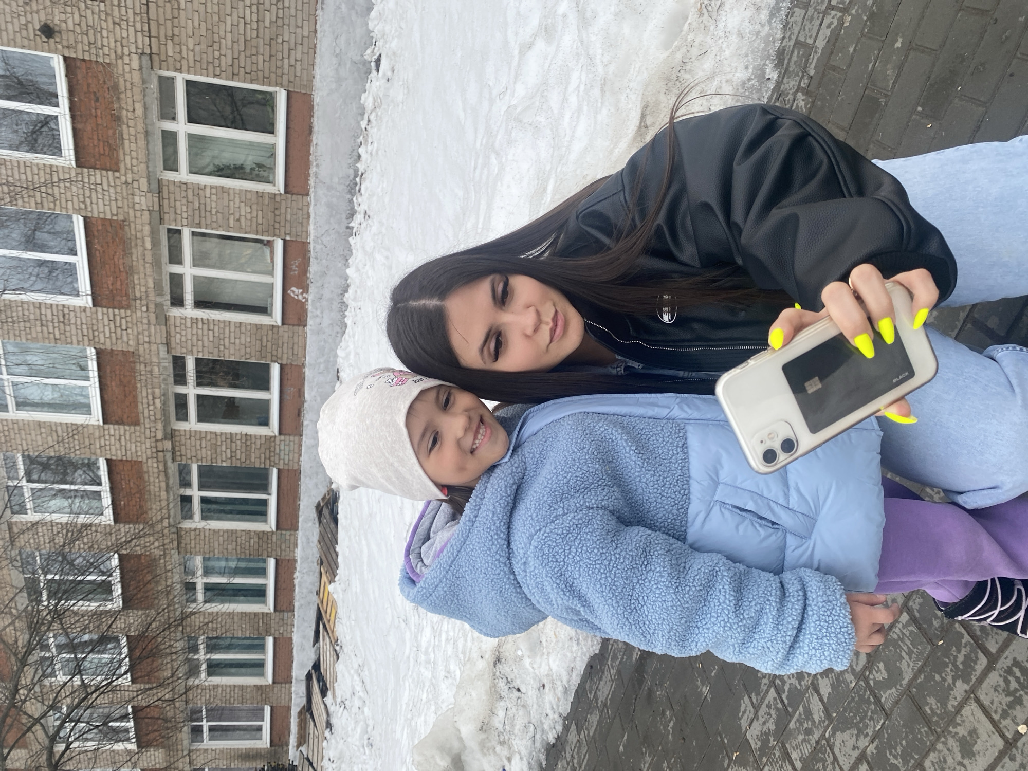 This outdoor image, captured in the city of Chaikovsky, Russia, depicts two females positioned on a wet paved surface. An adult female, appearing to be in her late teens or early twenties, with long dark hair, is crouching slightly while holding a light-colored smartphone with a dual camera array in her right hand, displaying neon yellow fingernails. Her attention is directed towards the phone's screen. She is dressed in a black faux leather jacket and light blue jeans. To her left and slightly in front stands a smiling child, approximately 6-8 years old, wearing a white knitted beanie, a light blue textured hooded jacket over a lighter blue puffer jacket, purple trousers, and black shoes with white laces.
The immediate ground beneath the subjects is a dark grey, wet brick or paver pathway. Behind the subjects, a substantial layer of dirty, melting snow covers the ground. Further back, a low, light-colored concrete or stucco foundation is visible, with wooden pallets or debris piled against it. The background consists of a multi-story building constructed from light brown brick, featuring reddish-brown horizontal bands separating floors. Numerous white-framed rectangular windows are visible, some reflecting bare tree branches. The scene suggests a cold-weather environment, likely winter or early spring.