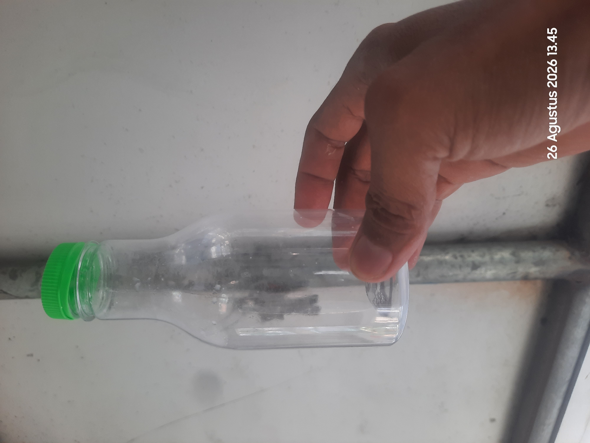 A clear plastic bottle with a green cap is held in a person's hand. The bottle is empty and appears to be a single-serving beverage container. The background is a light-colored, textured surface, possibly a wall or metal sheeting. A portion of a gray metal pipe is visible vertically behind the bottle. The bottom right corner of the image displays a timestamp: "26 Agustus 2026 13.45". The scene appears to be outdoors or in a non-residential setting, given the visible piping and simple background. The lighting suggests daytime.