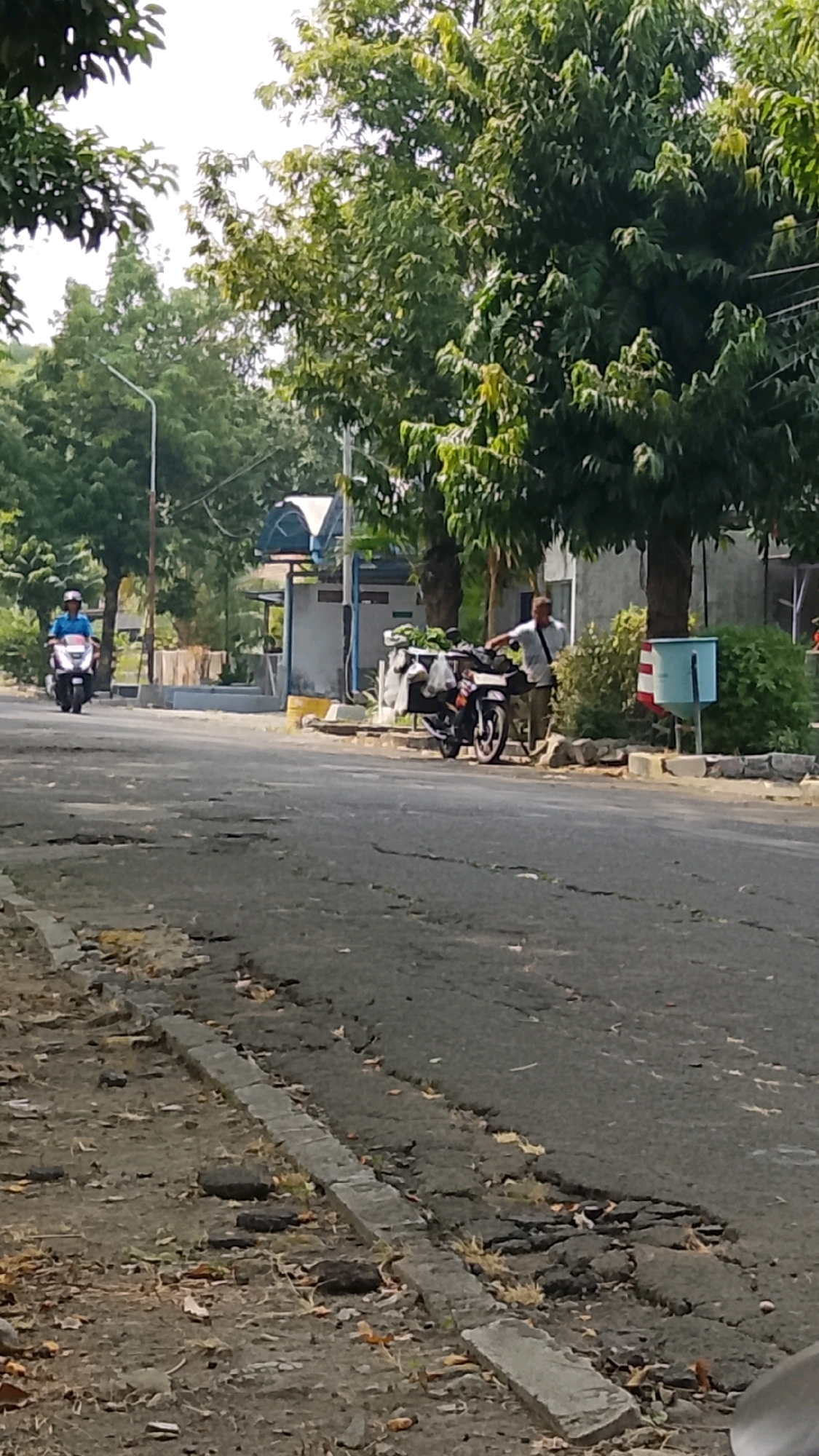 The image depicts a street scene in Gapurosukolilo, Indonesia. In the foreground, the asphalt road is cracked and uneven, with debris scattered along the side. On the right side of the road, a man is standing next to a motorcycle that is loaded with bags, some containing green produce. A blue trash receptacle with red and white stripes is situated behind the motorcycle. In the background, trees with dense green foliage line the street. Further down the road, a person on a white and black motorcycle is riding away from the camera. In the distance, a building with a light-colored roof and blue trim is visible. The sky is bright, suggesting daylight.