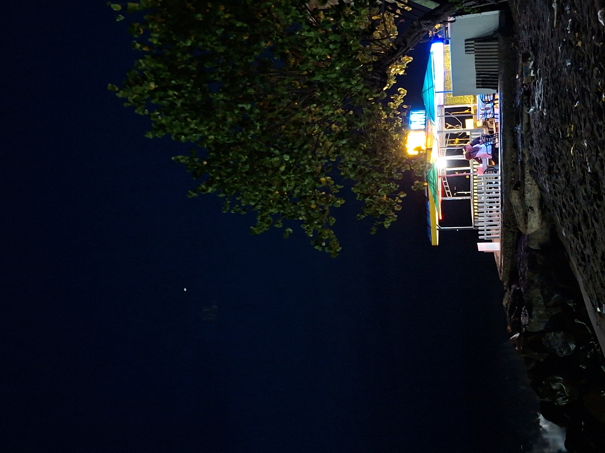 A nighttime scene depicts a coastal establishment in Lhokseumawe, Indonesia, characterized by artificial lighting and natural elements. In the foreground, dark, coarse sand is visible, interspersed with scattered debris. Beyond the sand, a rocky shoreline is present, with waves crashing against the rocks on the left side of the frame, creating a white foam.
Dominating the mid-ground is a small, open-air structure with a multi-colored roof, featuring yellow, green, and blue awnings. This structure appears to be a food or drink stall, or a small restaurant. It is illuminated by bright, colorful lights, including a prominent yellow light source above the central area and blue lights on the right. A white picket fence encloses a portion of the area. Inside, two individuals are seated at a table, one in a white top and the other in dark clothing. Several empty chairs and tables are also visible.
To the right of the structure, a large tree with dense green foliage extends into the frame from the right edge. The leaves are partially illuminated by the lights from the establishment.
The background is a dark, clear night sky, with a faint, small white object, possibly the moon or a distant light source, visible in the upper left quadrant. The overall atmosphere is dimly lit, with the artificial lights of the establishment providing the primary illumination.