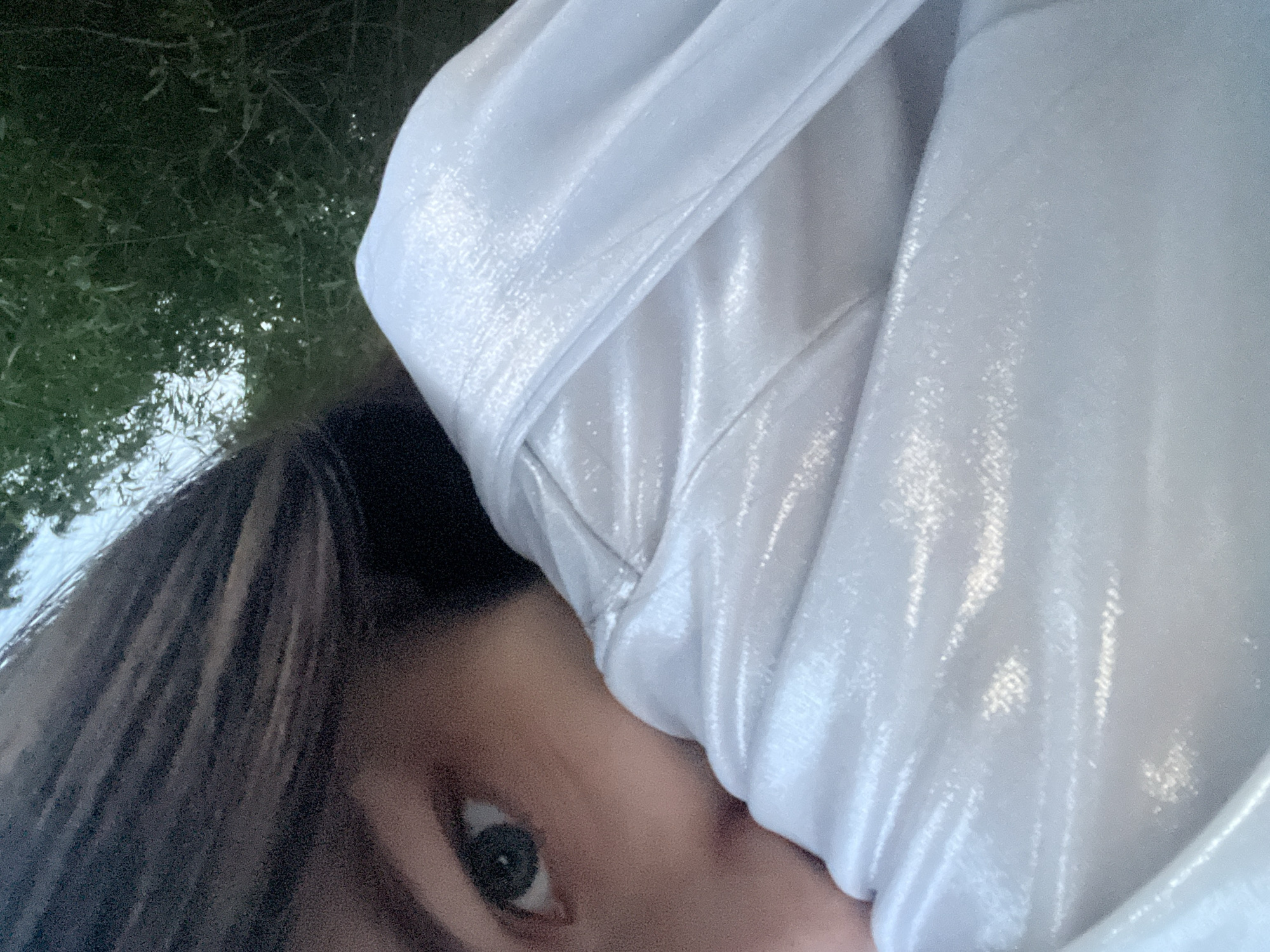 A close-up, diagonal shot captures a person partially obscured by a shimmery, light-colored fabric. The person's left eye, a portion of the nose, and cheek are visible. The eye features a dark pupil and a lighter-colored iris, appearing green or hazel. Dark brown hair is visible along the top and left edges of the frame. The fabric, which appears white or very light silver with a metallic sheen, is draped around the person's lower face, creating soft folds and reflecting ambient light. The heavily blurred background consists of green foliage, indicating an outdoor or garden setting. There is no discernible information within the image to identify the specific location as Sinyavino, Russia, or any other particular city.