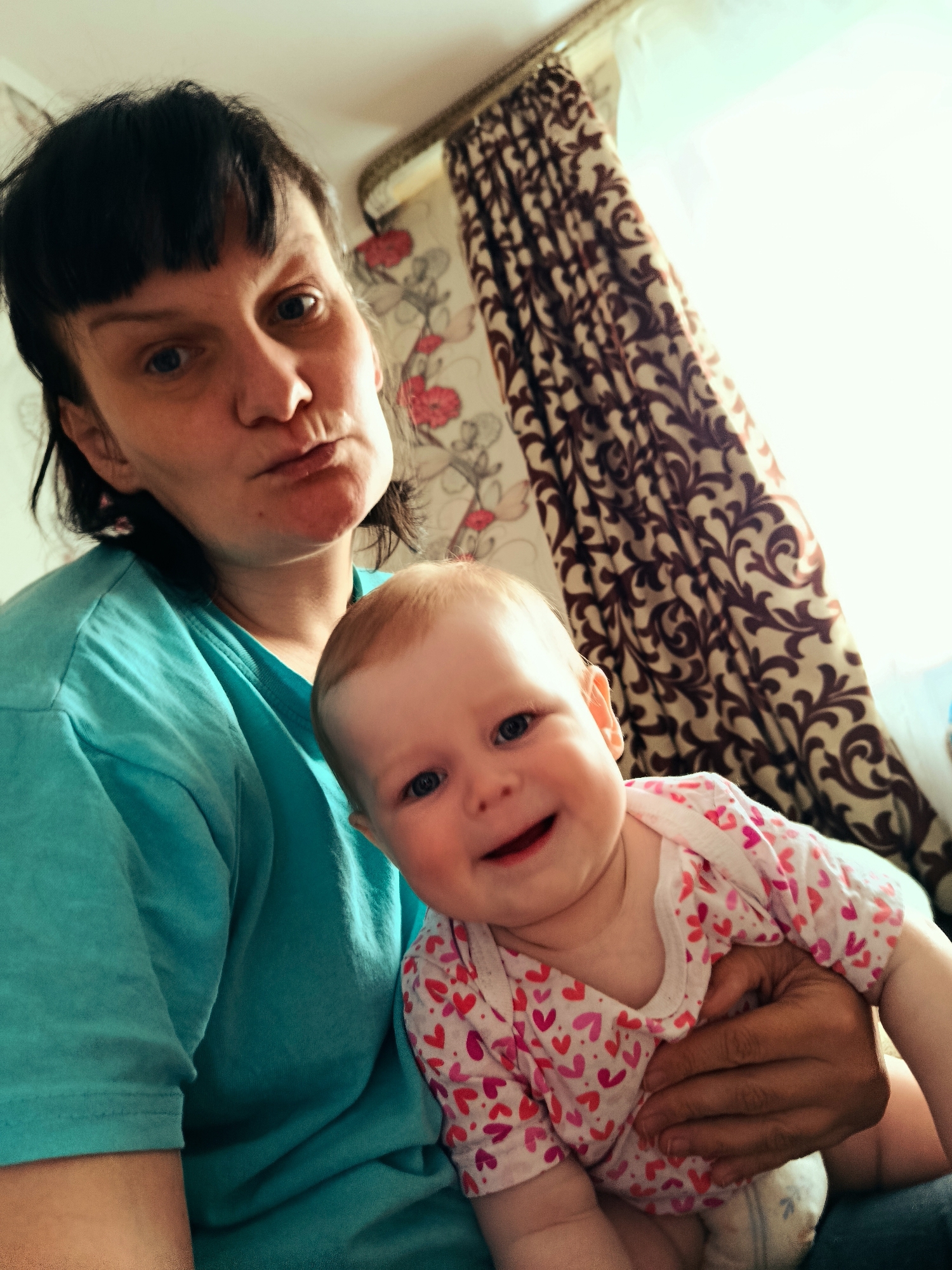 A woman is holding a baby. The woman has dark hair and a blue shirt. The baby is wearing a white shirt with red hearts. The baby is smiling. The background includes patterned curtains. The location is unknown.