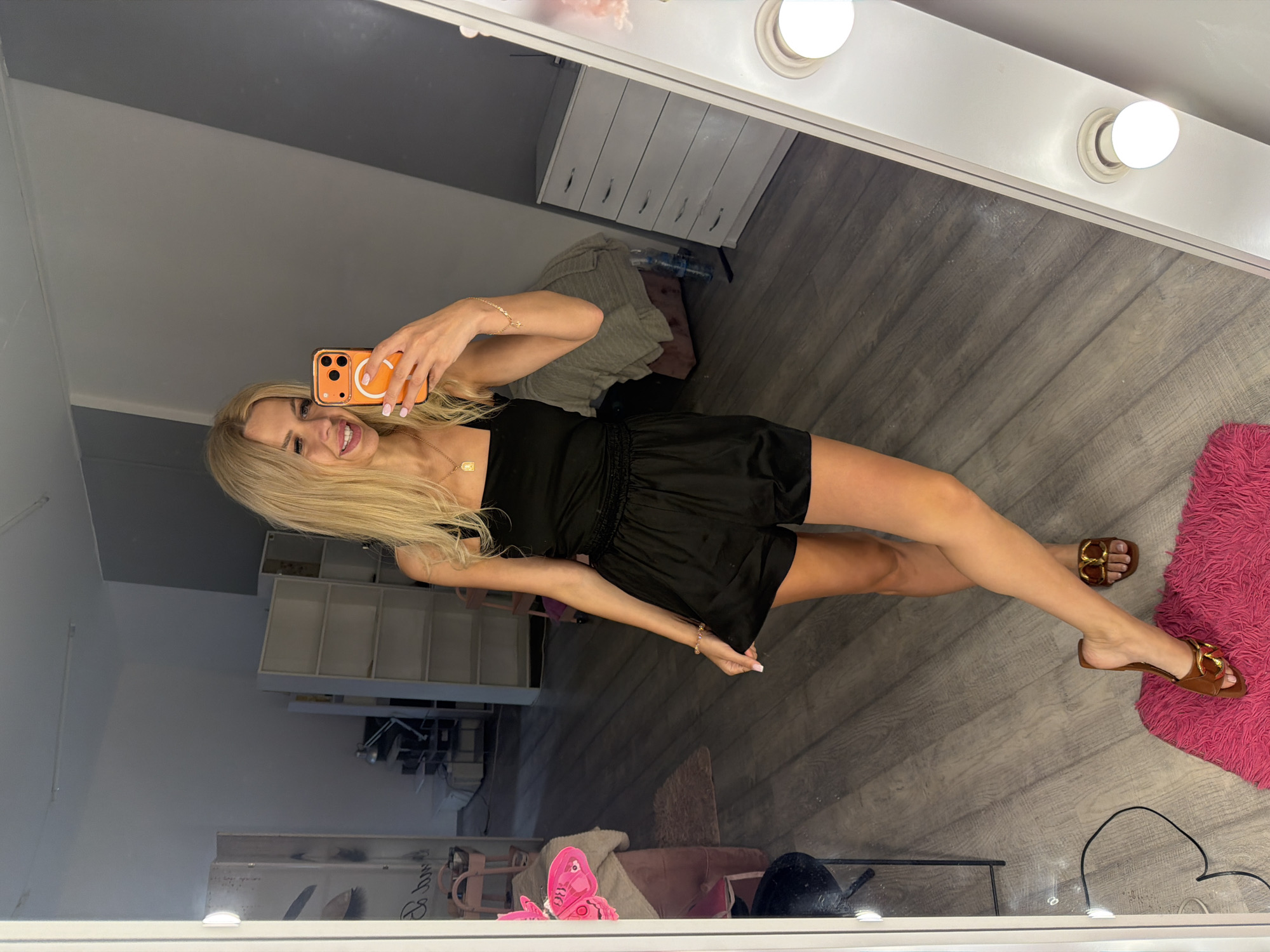 A woman with blonde hair is taking a selfie in a mirror. She is wearing a black romper and brown sandals. She is smiling and holding an orange phone. The background includes a dresser, shelves, and a pink rug. The room appears to be a dressing room or vanity area, suggested by the lights around the mirror. The floor is made of wood-look laminate. The overall mood is casual and confident. The time of day is not discernible, but the lighting suggests an indoor setting. There is no visible text in the image.