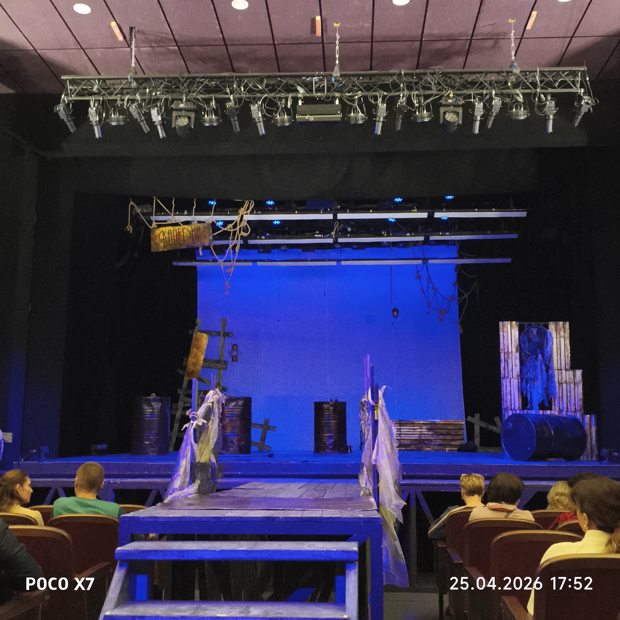 Scene: Theater performance in Chaikovsky, Russia.
Setting: A theater stage is set with a blue backdrop, barrels, wooden structures, and props resembling a post-apocalyptic or dilapidated setting. Audience members are seated in the foreground.
Objects: Stage lights are visible overhead. Props include barrels, wooden platforms, a ladder, and a sign with illegible Cyrillic writing.
People: Audience members are seated, their backs mostly facing the camera.
Actions: A performance is implied by the set design and lighting.
Environment: Indoor theater setting. The timestamp indicates April 25, 2026, at 17:52.