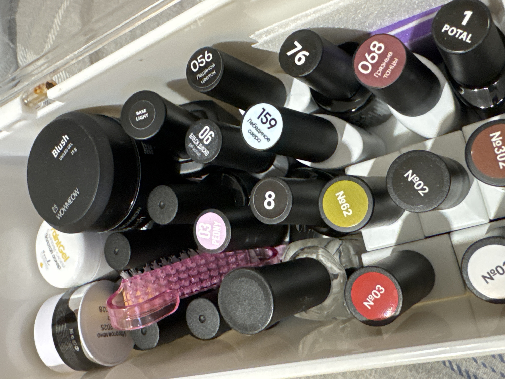 The image shows an overhead view of a white plastic organizer filled with various nail polish and gel products. The organizer is positioned on a grey and white checkered surface, likely a textile. The products include small jars with black lids, some with labels indicating "Blush UV/LED GEL 15g" and "WOMMEOW". There are also numerous bottles with black caps, each featuring a circular label on top with numbers and text in Russian. These labels include "03 PEONY", "8", "056 ЛЕДЯНОЙ ЦВЕТОК", "90", "159 ЛЕДЯНОЕ ОЗЕРО", "76", "№03", "№62", "№02", "№01", "№302", "068 ГРЯЗНЫЕ ТАНЦЫ", and "1 POTAL". A pink, textured grooming tool, possibly a nail brush, is also visible among the bottles. A small glass vial with a liquid and a metal cap is present in the upper right corner. The scene is indoors, and there are no people visible. The items appear to be new or in good condition. The location is identified as Яромінскі сельскі Савет, Belarus.
