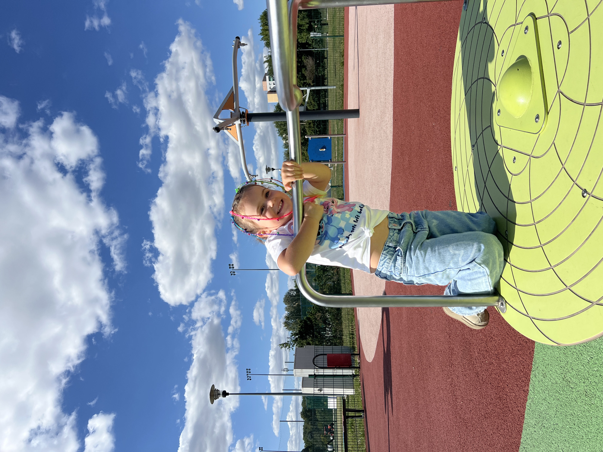 A young girl with braided hair adorned with colorful ribbons kneels on a bright green, circular spinning platform at a playground. She wears a white t-shirt with a cartoon print and light blue jeans. Her hands grip a silver metal railing. The playground has a red and pink rubberized surface. In the background, there are streetlights, a fence, trees, and a blue sky with fluffy white clouds. The image appears to be taken at a modern outdoor public recreational area.