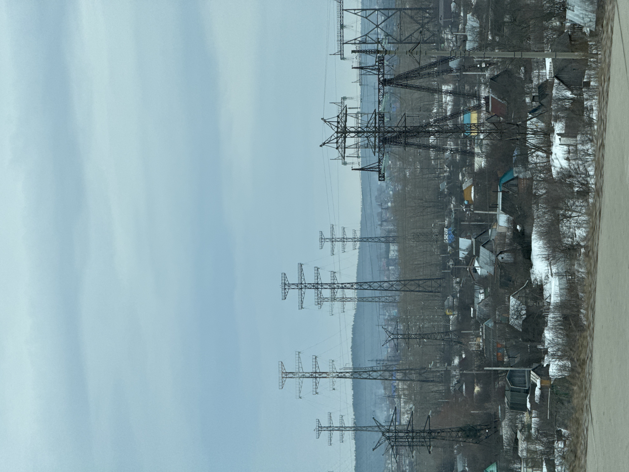 The image depicts a power line passing through a residential area near Chaikovsky, Russia, likely taken during winter. Steel lattice power towers are lined up vertically on the left side of the frame. A collection of houses with a dusting of snow and bare trees are seen on the right. The sky is a light blue-grey with subtle cloud variations, typical of overcast weather.