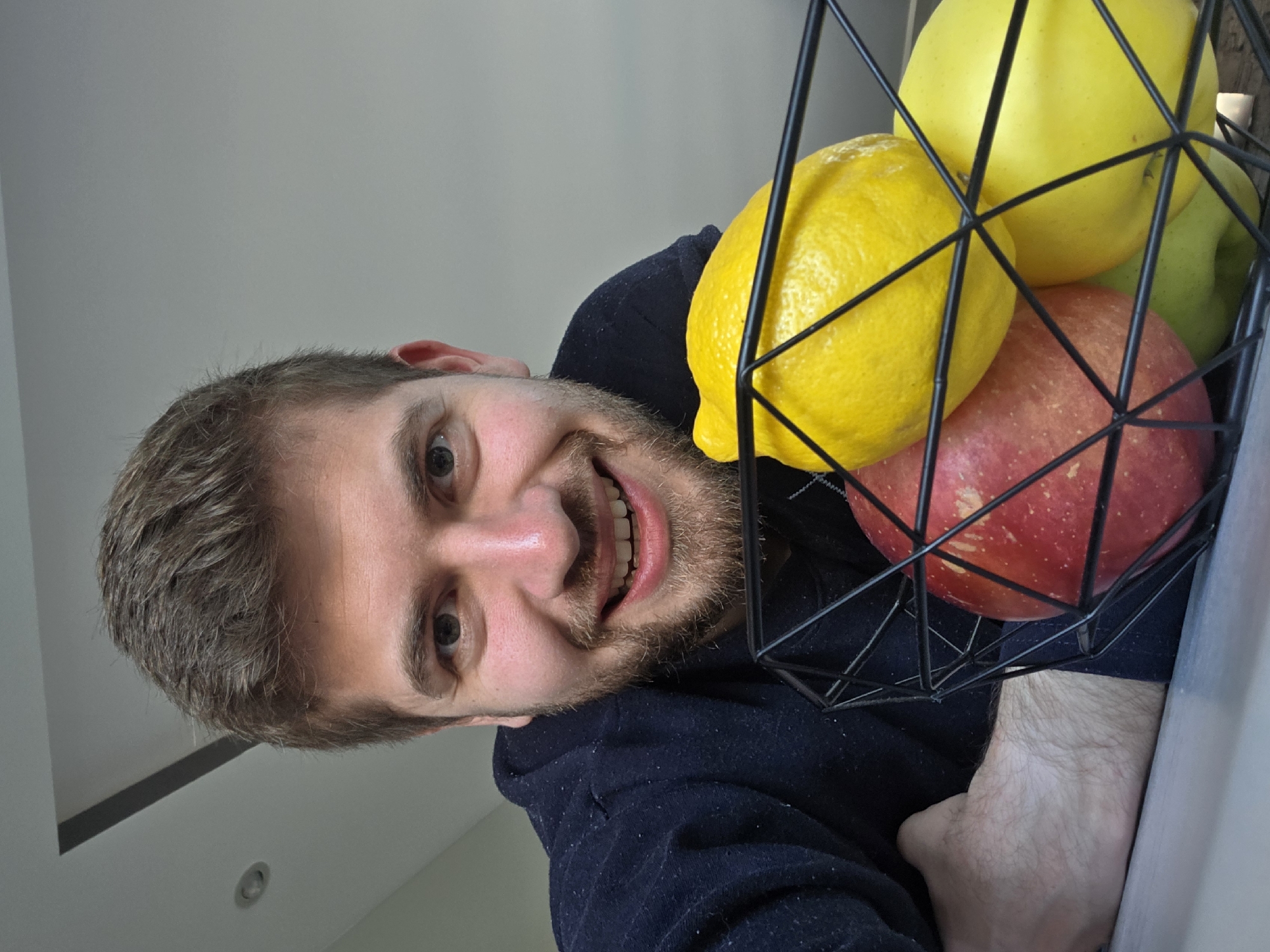 A man with brown hair and a beard is smiling at the camera. Next to him is a black wire basket filled with fruit. The fruit consists of two lemons, a red apple, and a green apple. The background is a neutral white wall. No indication of Yagodovo, Bulgaria.