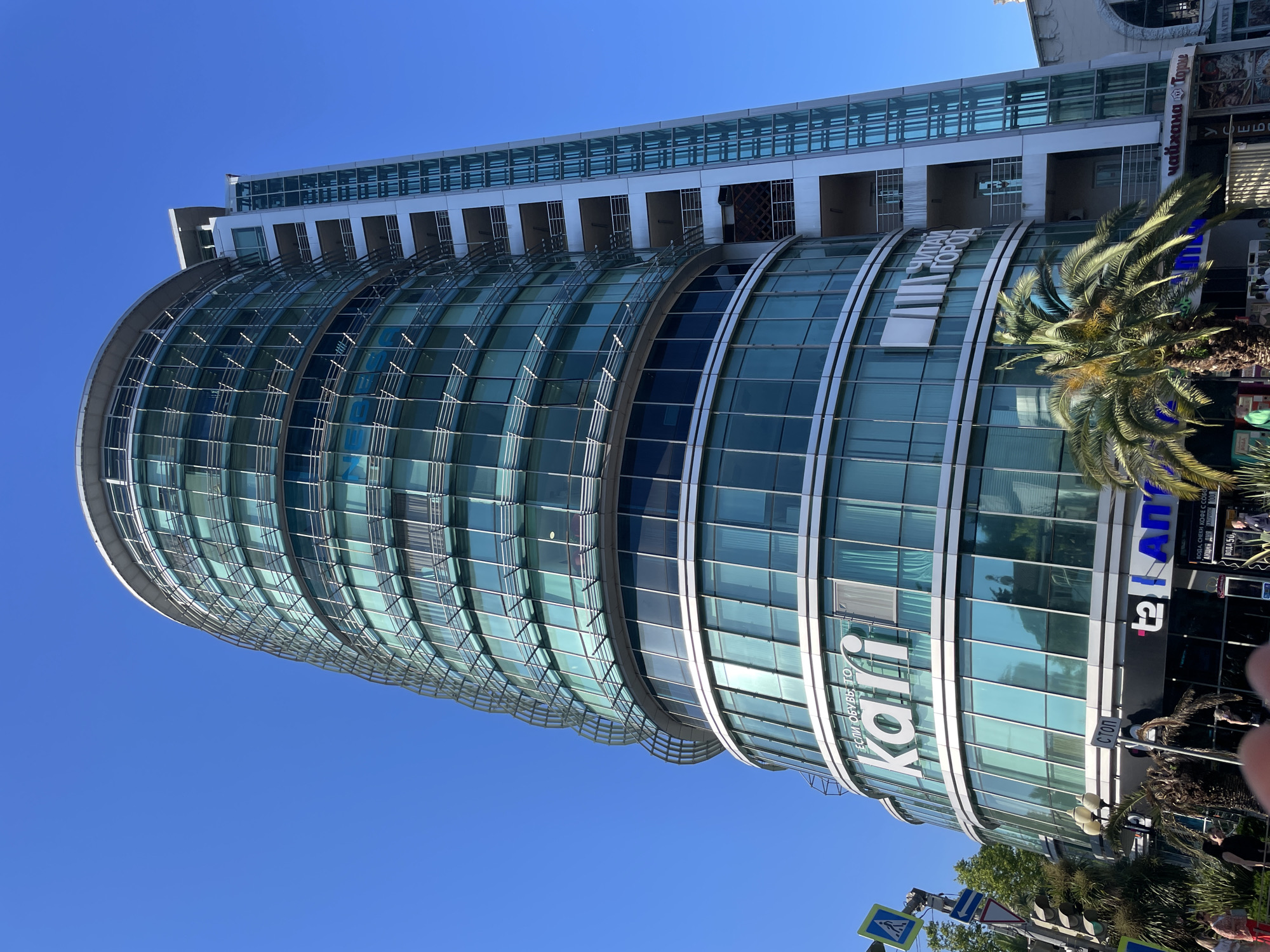 This is a medium-angle, outdoor, full-shot photograph of a modern, curved glass building under a clear blue sky. The building is tall with multiple floors, characterized by its smooth, reflective glass facade with horizontal banding. A palm tree in the foreground partially obscures the lower portion of the building on the right side. Traffic lights and street signs are visible at the bottom of the image, suggesting an urban setting.
Visible text on the building includes: "ЕСЛИ ОБУВЬ, ТО" and "kari" in large white letters on the glass, and smaller signs that read "ЧИТАЙ", "Город", "Стоп", "ΑΠ", "АКЦИЯ ДНЯ", "ВОДА, СНЕКИ КОФЕ С СОБОЙ", "ВОДА 50", and "чайхана Торке". The presence of Cyrillic script, the "kari" brand (a Russian footwear retailer), and palm trees are strong indicators of the location being Sochi, Russia.
The lighting suggests it is daytime, likely mid-morning or afternoon, given the bright sunlight and clear sky. The overall scene is bustling with commercial activity, as indicated by the storefronts and signage. There are no prominent people or specific activities depicted, but the setting suggests a lively urban environment.