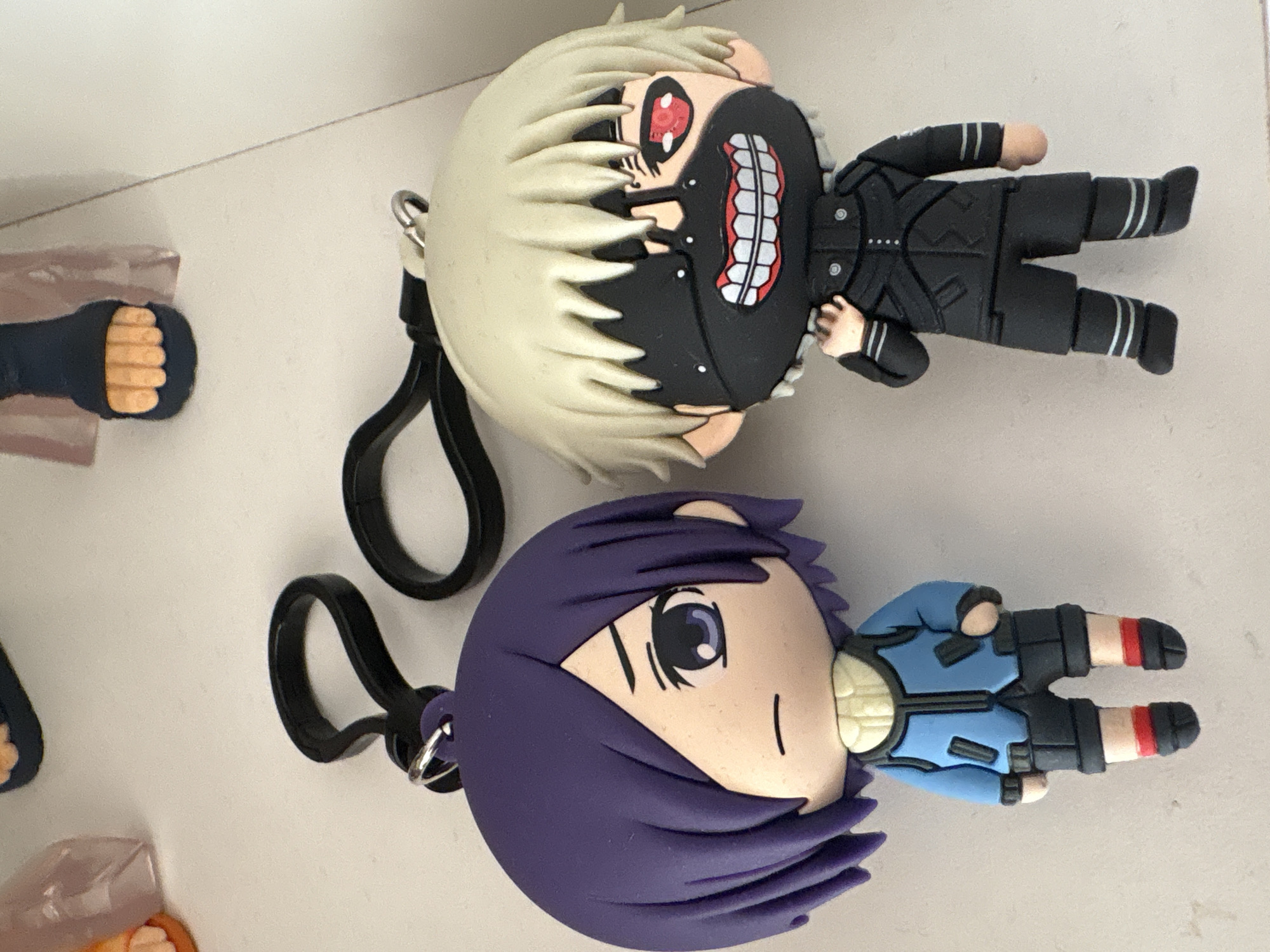 Two cartoon character keychains, presumably made of rubber, are lying on a flat, light-colored surface. The top keychain portrays a character with white spiky hair, a black mask covering part of the face revealing a red eye and teeth, and wearing a black suit. The bottom keychain portrays a character with purple hair covering part of the face, showing one eye, and wearing a blue suit. Both keychains have black clips attached. Fragments of other similar items are visible on the left side of the image. The location in Plovdiv, Bulgaria cannot be determined from the image.