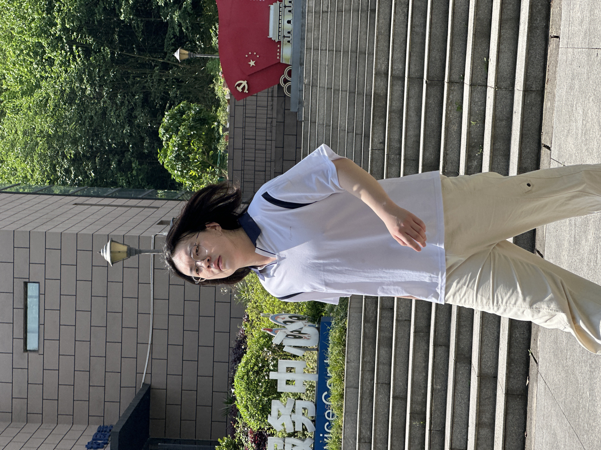 An outdoor, sunlit, full-body portrait captures an adult with long dark hair and glasses, wearing a white short-sleeved polo shirt with a dark blue collar and trim, and light beige pants. They stand with a neutral expression, looking directly at the camera, positioned on level ground next to a concrete stairway.
In the background, a light-colored brick building is partially visible to the person's left, featuring a small rectangular window and a classic-style street lamp mounted on its wall. Lush green bushes and trees frame the scene, appearing both in the foreground near the person and extending into the background.
A notable red sign or emblem is visible above the stairs in the background. It prominently displays a golden hammer and sickle symbol along with five golden stars, indicative of the Communist Party of China. Indistinct Chinese characters are also visible on this red sign, likely part of a building name or official motto. Below the person's eye level, amidst the foliage, a blue sign with white text is partially visible, reading "中心" (zhōngxīn, center) and "Service Ce" (likely part of "Service Centre").
The concrete stairway, with numerous steps, ascends to the right side of the image. Strong shadows cast by the person and on the stairs suggest bright daylight, indicating a sunny day, likely during midday or afternoon. The setting appears to be an urban or campus environment in Jiangjin, China.