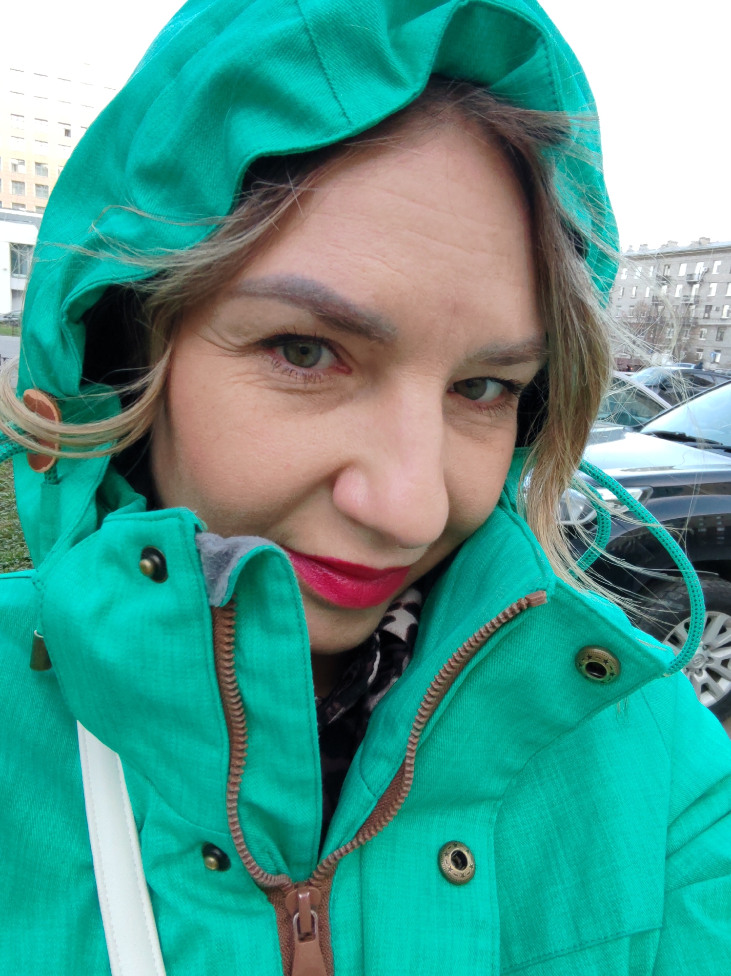 A selfie taken in the city of Saint Petersburg, Russia, features a woman wearing a bright green hooded jacket. The hood is up, partially covering her blonde hair. She has green eyes and is wearing red lipstick. Her skin shows some wrinkles around her eyes. The zipper of her jacket is partially open, revealing a glimpse of a scarf underneath. In the background, there are several cars and apartment buildings, suggesting an urban setting. The sky is overcast.