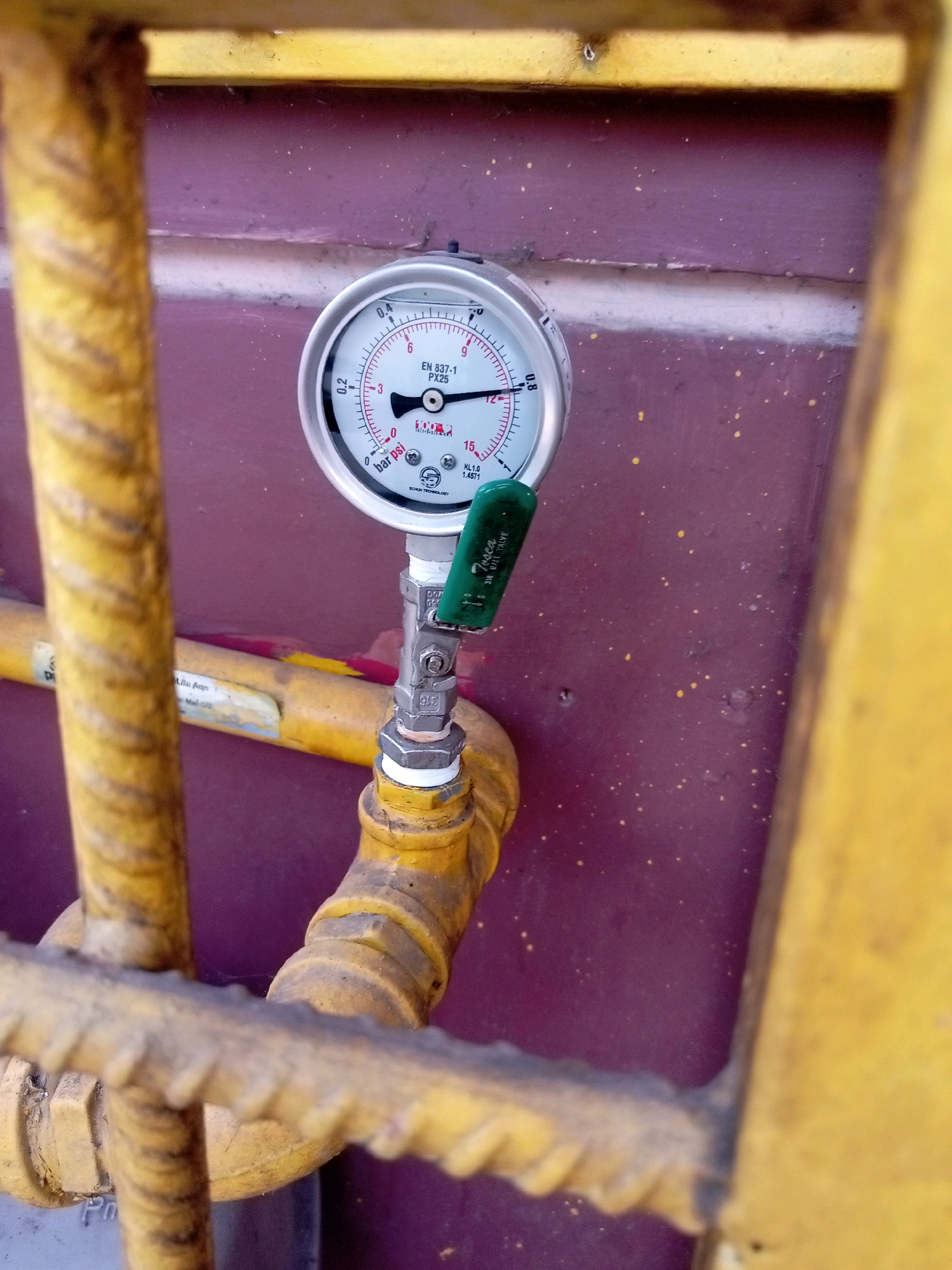 A close-up, eye-level shot captures a pressure gauge and valve on a yellow pipe, positioned in front of a textured, purple wall. The gauge, mounted vertically, has a white face with black markings and a red needle. The valve, attached to the gauge, has a green lever. The yellow pipe, with visible threaded connections and some wear, extends horizontally and curves downwards. The background wall has a rough texture and is speckled with small yellow dots. The foreground is dominated by blurred yellow bars, suggesting a metal grate or railing. The overall scene appears to be located outdoors, possibly in an industrial or utility area, with the weathered appearance of the equipment and the outdoor setting. The image does not contain any people, actions, or specific interactions. It is a static capture of a piece of equipment. The scene does not provide any direct indicators of its location within Batam, Indonesia, such as signs or landmarks.