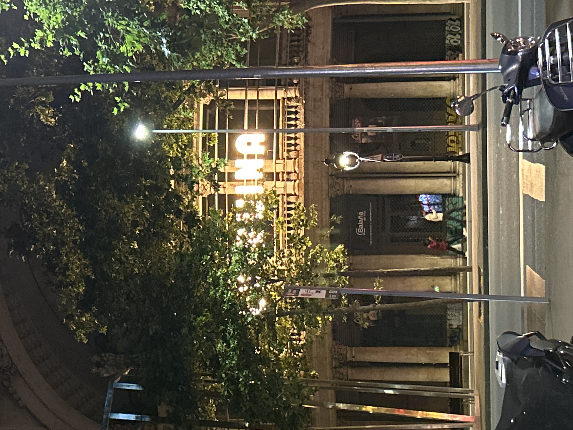 Captured at night in Barcelona, Spain, the image presents an urban scene viewed from a slightly angled perspective. A building with illuminated lettering reading "TINA" is centrally placed, partially obscured by foliage. Street lamps and trees dominate the foreground. To the right, motorcycles are parked along the street's edge. The background reveals parts of the building facade, including columns. People are visible in the building's interior. The scene has a mix of natural and artificial lighting creating contrasts and shadows.