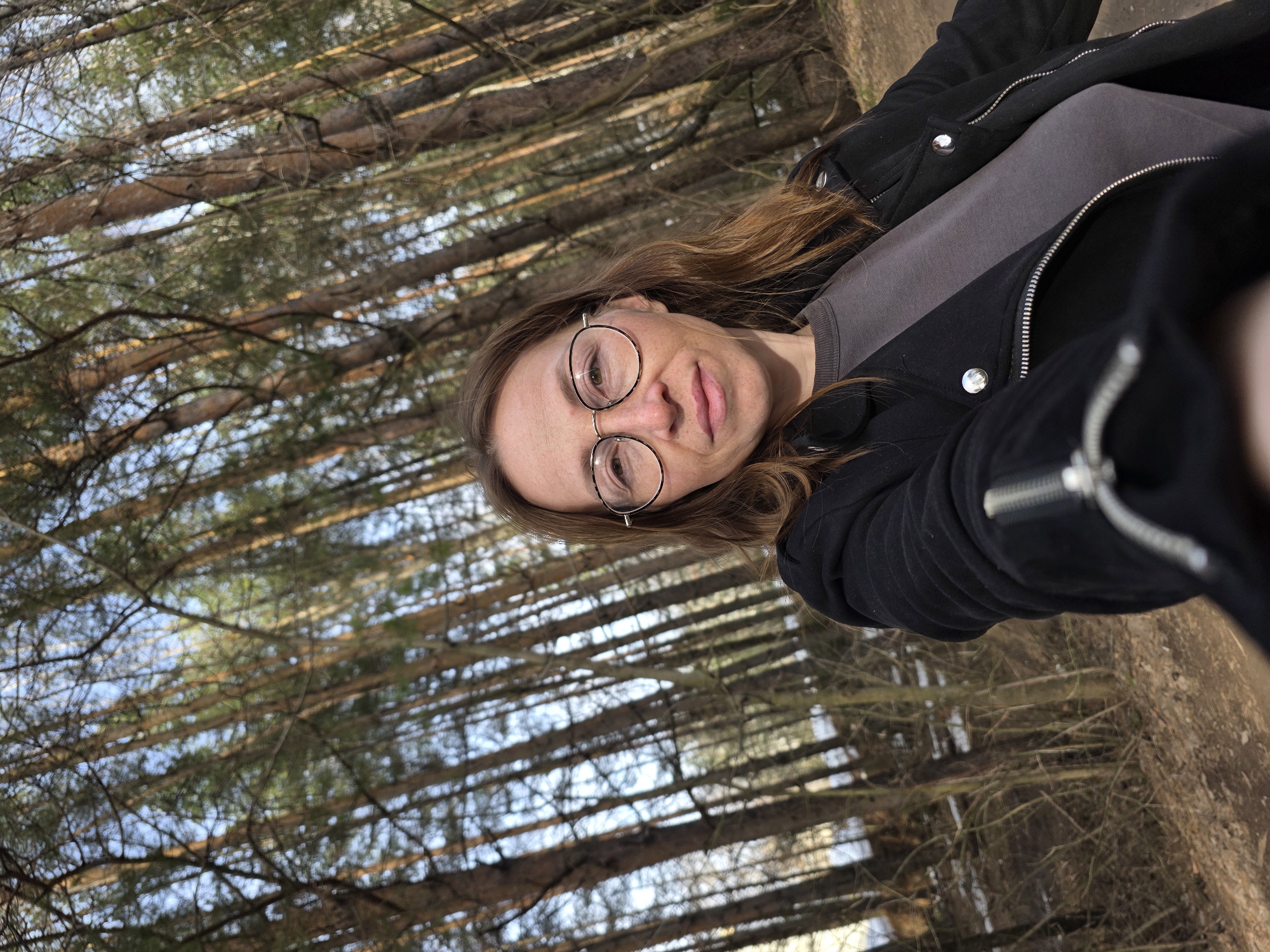 A woman with long brown hair and glasses is taking a selfie in a forest. She is wearing a gray shirt and a black jacket. The forest has tall, straight trees with brown bark and green needles. The sky is visible through the trees and is blue. The location is likely in a wooded area near Chaikovsky, Russia. The ground is covered with brown leaves and dirt.