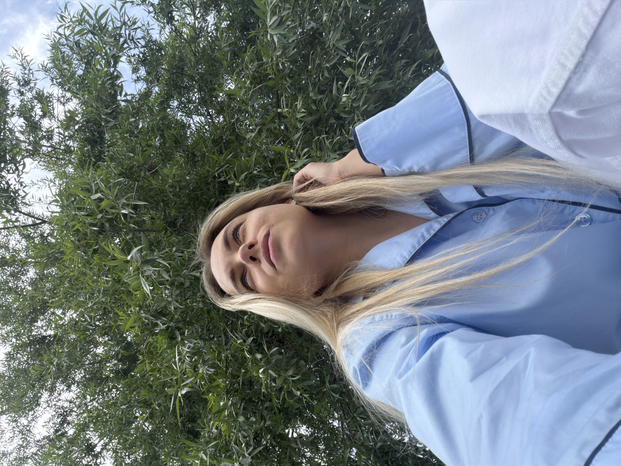 A close-up outdoor portrait features a young woman with long, straight blonde hair. She is captured from a low angle, gazing upwards and slightly to her left, with a calm, neutral expression. Her eyes are light-colored, and her lips are full. She wears a light blue button-up shirt with contrasting dark blue or black piping along the collar, cuffs, and front placket. One hand is visible near her right ear, gently touching her hair.
The background is dominated by dense, vibrant green foliage, likely from a tree with slender leaves, possibly a willow. Patches of bright, partly cloudy sky are visible through the leaves, indicating daytime. A small portion of a white garment or object is visible in the upper right corner of the frame. The natural lighting suggests bright daylight. The scene conveys a serene, outdoor moment, likely in a park or natural setting within Votkinsk, Russia. There is no visible text in the image.