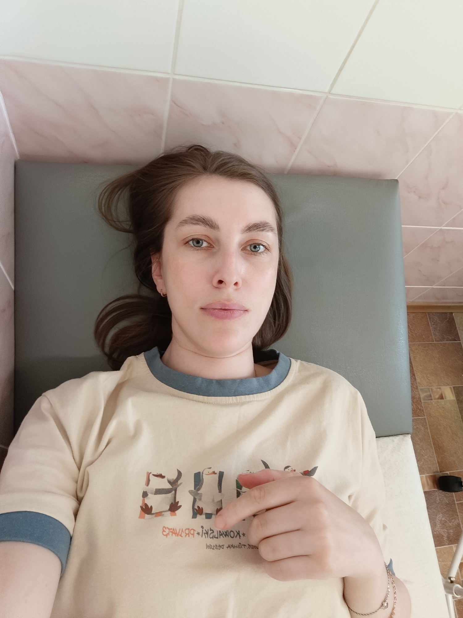A woman with light brown hair and blue eyes is lying on a grey padded surface, possibly a medical exam table. She is wearing a beige t-shirt with blue trim and a graphic print. Her right hand is slightly raised. The background consists of tiled walls and flooring. It's impossible to verify the location without additional information or visual clues within the photo.