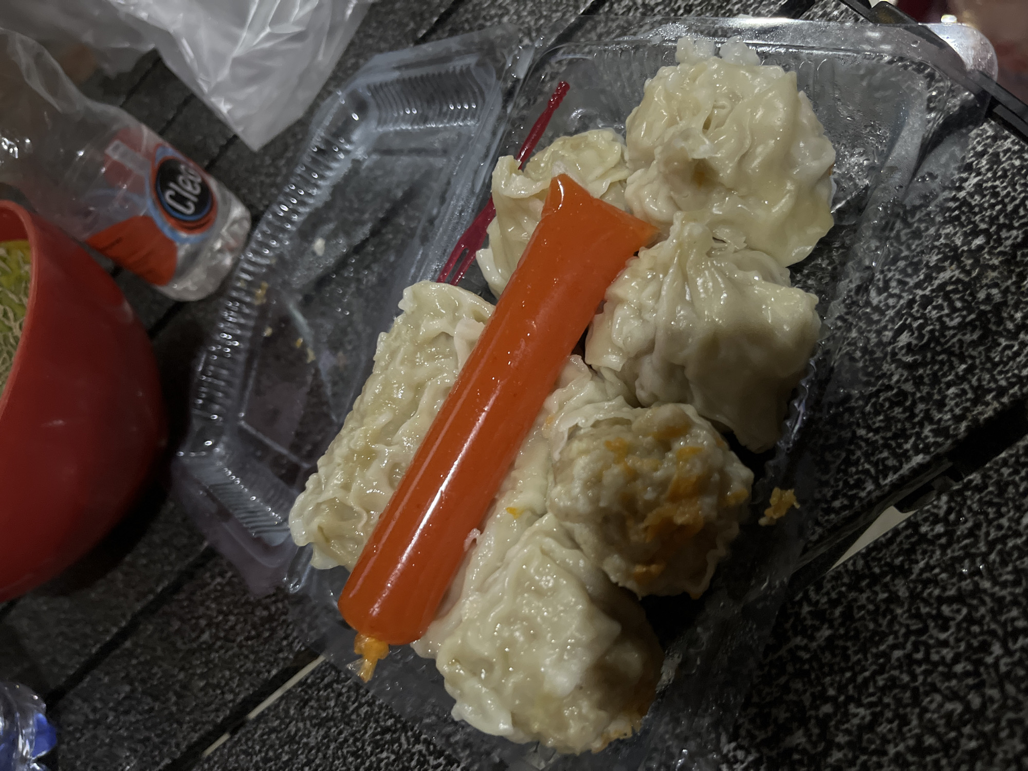 In Karangrandu, Indonesia, a close-up shot reveals a clear plastic container filled with steamed dim sum. One piece of dim sum is skewered with an orange processed food stick, possibly imitation crab meat. A red plastic fork is partially visible nestled amongst the dim sum. The container rests on a dark, textured surface, possibly a wooden table. In the background, a red bowl is visible, and next to it, a partially filled plastic bottle with a blue and white label that reads "Cleo." The overall lighting suggests an indoor or evening setting.