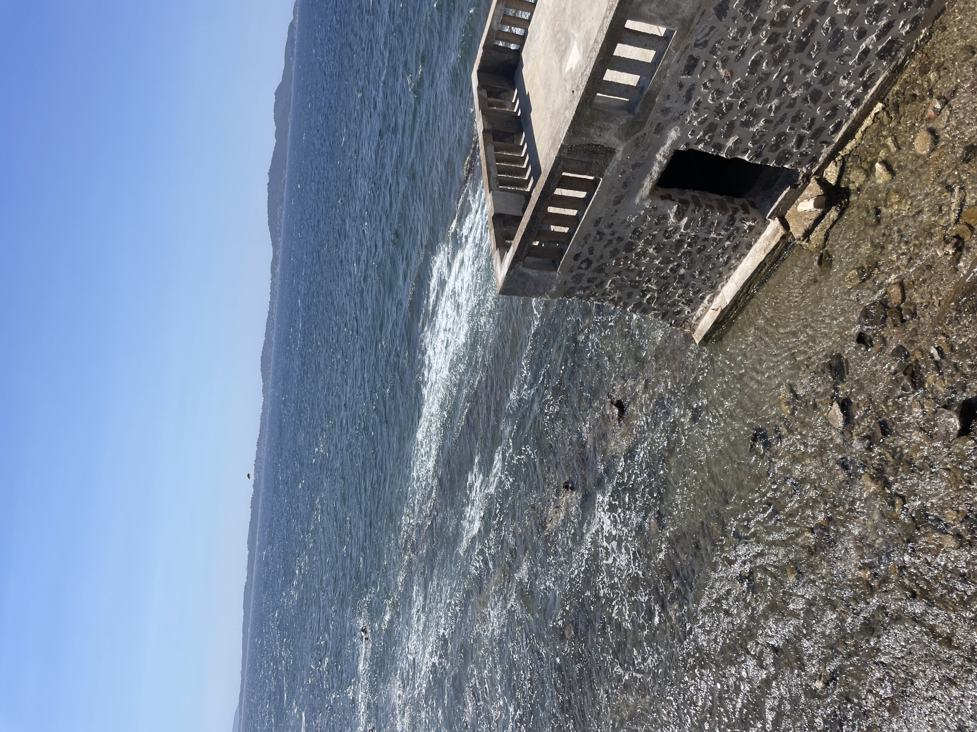 The image depicts a clear, sunny day with a bright blue sky over the Mediterranean Sea. In the foreground, a small stone building with a concrete balcony juts out over the water. The building has a rough stone facade and a doorway that appears to lead into darkness. The balcony has a concrete floor and a railing made of evenly spaced concrete posts. The water is choppy and appears shallow near the building, with rocks visible beneath the surface. Further out, the sea is a deeper blue, with distant hills or islands visible on the horizon. A small yellow object, possibly a parasail or kite, is visible in the far distance above the water. The scene is likely located in Carqueiranne, France, given the coastal setting and the architectural style of the building.