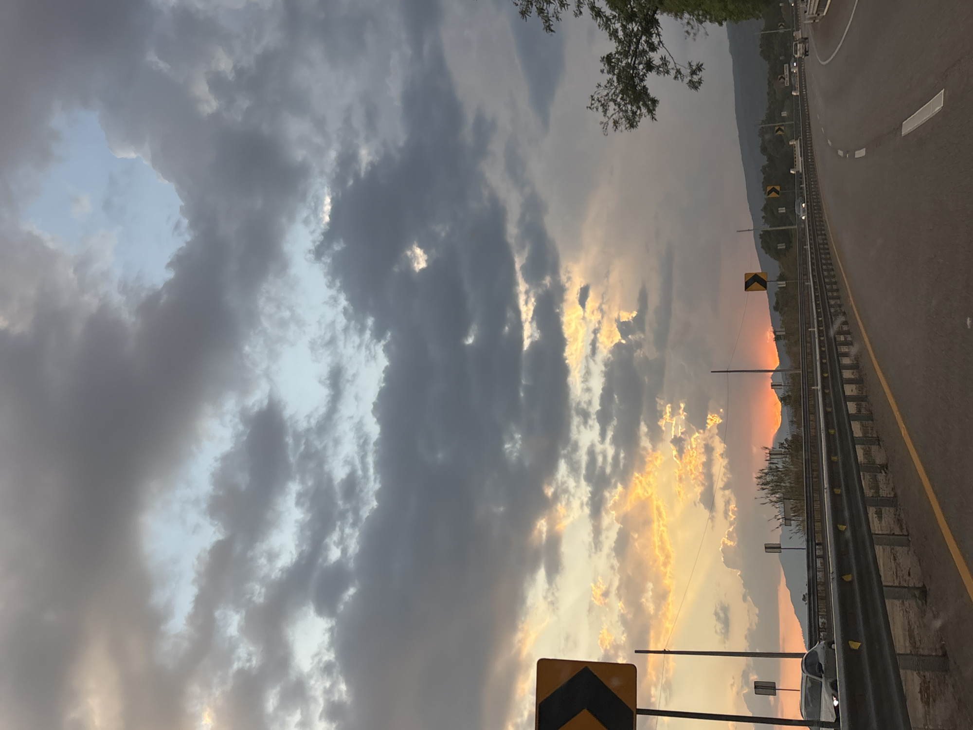 The image captures a sunset scene near a highway in Tepoztlán, Mexico. A cloudy sky dominates the view, with patches of blue and vibrant orange hues where the sun is setting. The highway is visible on the right side of the image, with metal guardrails, yellow lane markings, and road signs indicating curves. Mountains are silhouetted in the distance, further enhancing the scenic backdrop. A few cars are visible, but people are not present. The image is slightly tilted, adding a sense of dynamic perspective to the overall composition.
