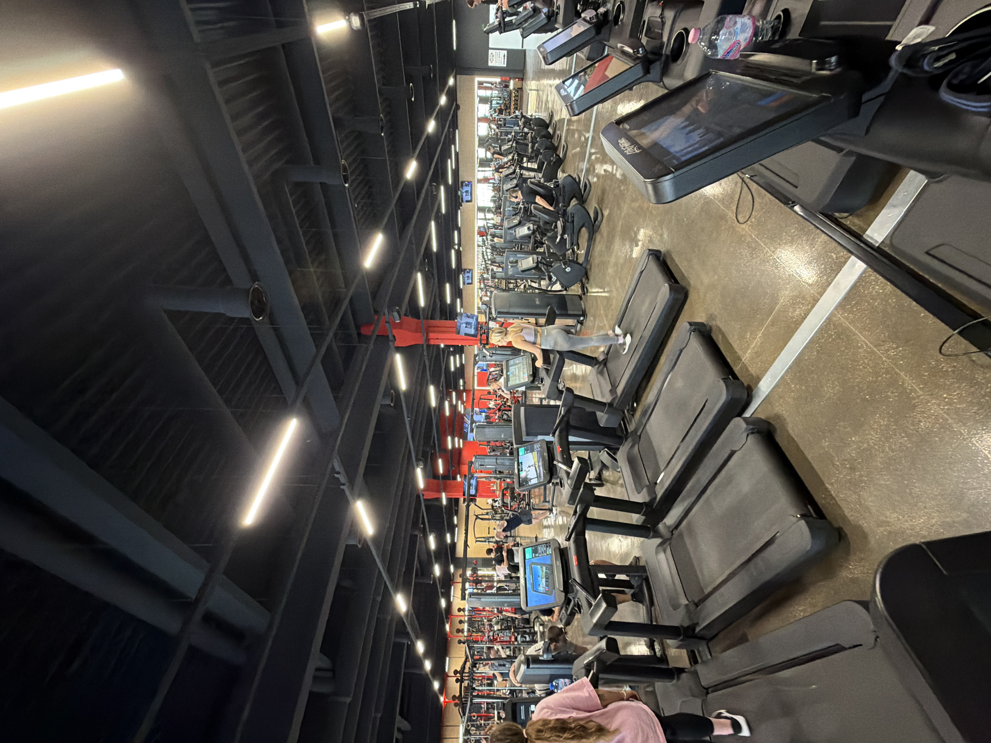 This image depicts a gym interior. The scene focuses on rows of treadmills, many of which are occupied by individuals. Additional gym equipment is visible further back in the frame. The interior is lit by overhead fluorescent lights, and a dark, industrial-style ceiling is present. The location and city mentioned are not ascertainable.