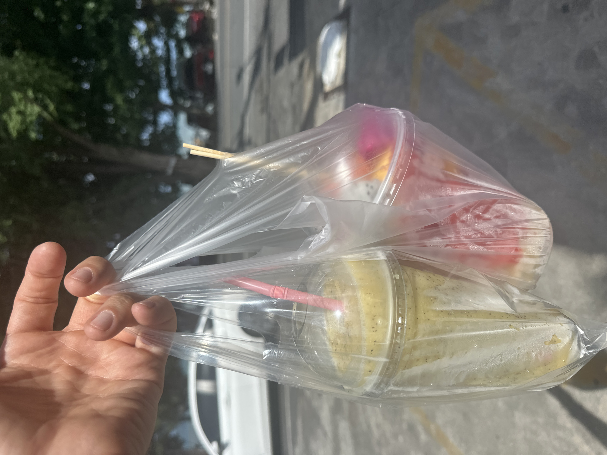 In Rawai, Thailand, a person's hand holds a clear plastic bag containing two drinks. One drink, on the left, is a pale yellow smoothie in a clear plastic cup with a pink striped straw. The other drink, on the right, is a pink and white fruit salad in a clear plastic cup, with wooden skewers protruding from it. The bag is translucent and slightly crinkled. In the background, a white car is visible on a street with trees and shadows cast on the asphalt. The scene suggests an outdoor setting, possibly a street-side vendor or a takeaway purchase.