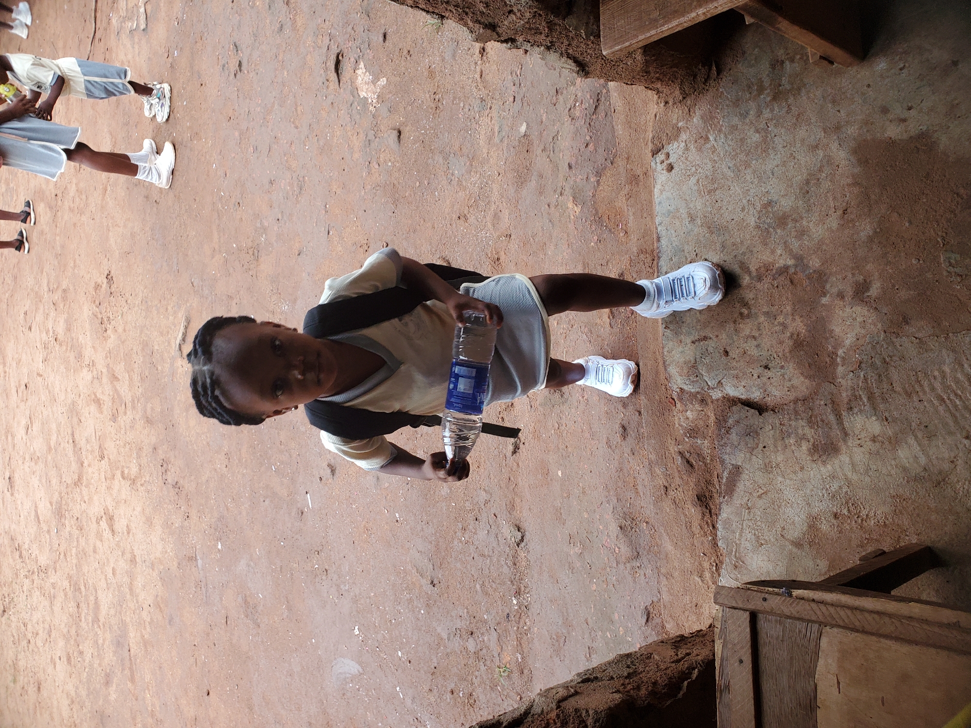 A child stands on a reddish-brown concrete or stone surface, holding a water bottle. The child wears a light-colored shirt, shorts, white sneakers, and a backpack. Other children are visible in the background, also in light-colored clothing and sneakers. The location appears to be outdoors, possibly a schoolyard. The background suggests a rough, possibly rural or less developed environment. The wooden structures in the top right and bottom right corners may be part of a building or furniture. The scene is in Ukwu-Abwa, Nigeria.
