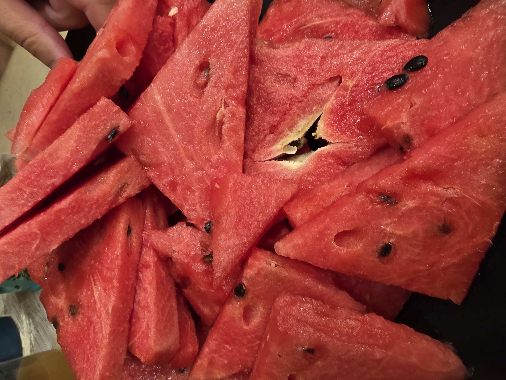 A close-up, overhead shot displays a pile of sliced watermelon on a black surface. The watermelon pieces are triangular and vary in size, with some exhibiting seeds. A hand is visible in the upper right corner of the image, suggesting someone is about to take a slice. The background is blurred but includes what appears to be a glass of amber liquid and a blue object, possibly another drink. The lighting is bright and focused on the watermelon, creating highlights on its juicy surface. The image does not provide any information about its location in Saint Petersburg, Russia.