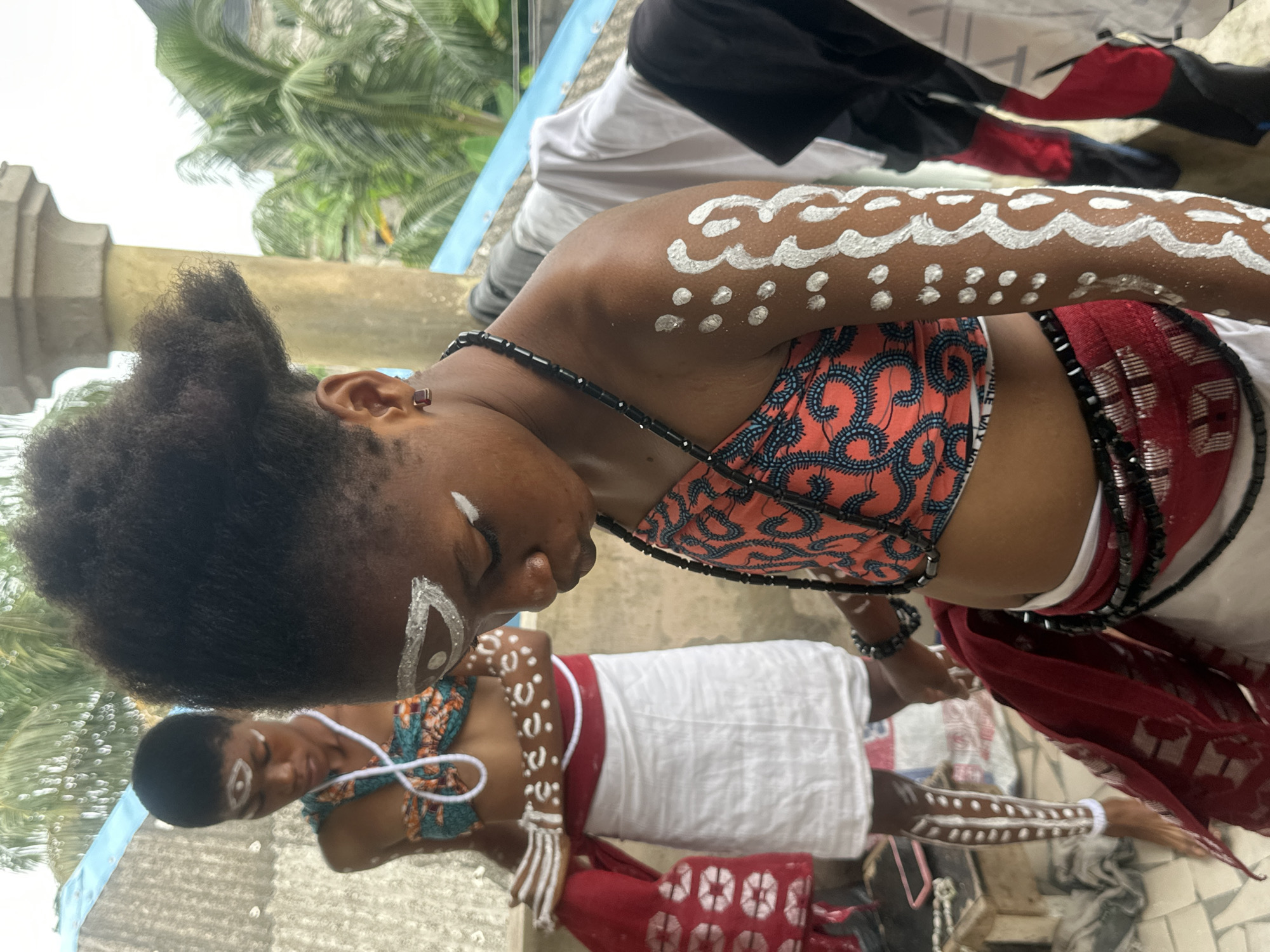 Two individuals, likely young girls, are adorned with white body paint in traditional patterns on their faces and limbs. The one in the foreground, facing right and looking downwards, wears a beaded necklace and a patterned bandeau top with matching beaded accents on her skirt. Her arm displays intricate white markings. Behind her, another individual, facing left and also looking downwards, is dressed in a similar bandeau top and a white skirt. Their legs are also painted with white designs. They appear to be participating in a cultural ceremony or ritual. The background suggests an outdoor setting with hints of roofing, possibly a dwelling or public space, with lush green palm trees visible, indicating a tropical environment. The presence of laundry hanging on a line to the right suggests a residential area. The overall scene evokes a sense of cultural tradition and spiritual practice within what appears to be a Nigerian urban or semi-urban environment.