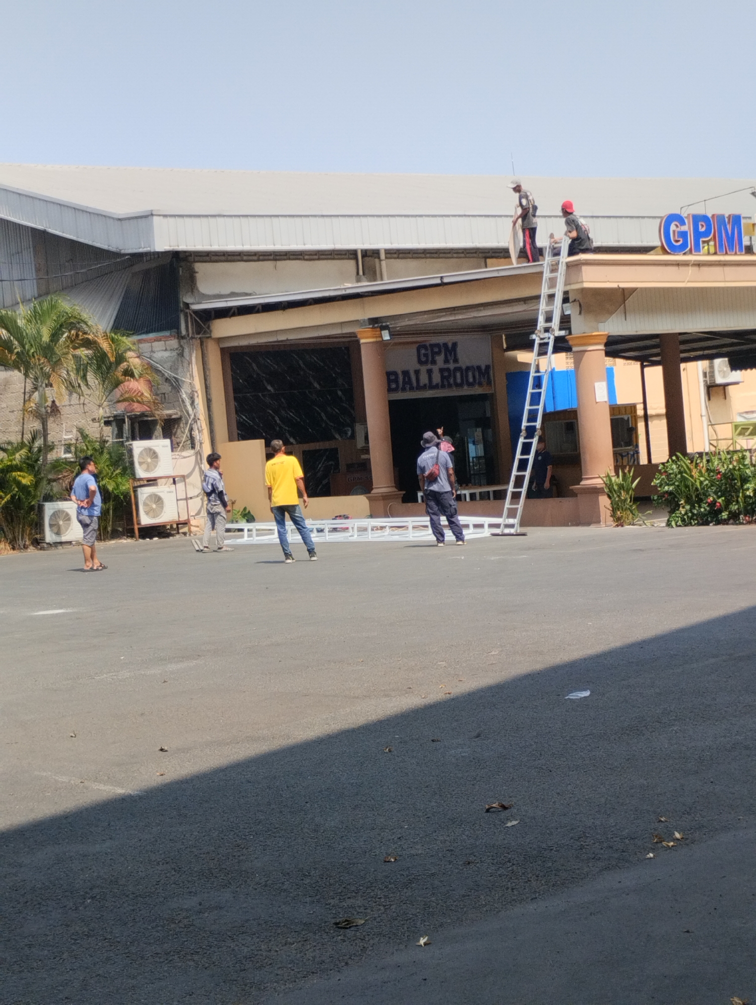 In Bekasi, Indonesia, a man in a red cap and another in a gray shirt are on a ladder near a building with a sign that reads "GPM BALLROOM." The man in the red cap is seated on the roof of the building, while the man in the gray shirt is standing on the ladder, holding what appears to be a piece of white material. Below the building, three men are standing on an asphalt surface. One man in a blue t-shirt and gray shorts is looking towards the building. Another man in a yellow t-shirt and blue jeans is standing with his back to the camera. A third man, wearing a gray shirt and pants, is also on the asphalt. Several white structures, possibly part of a temporary event setup, are laid out on the ground. The environment is bright and sunny, with a clear sky. Air conditioning units are visible on the side of the building.