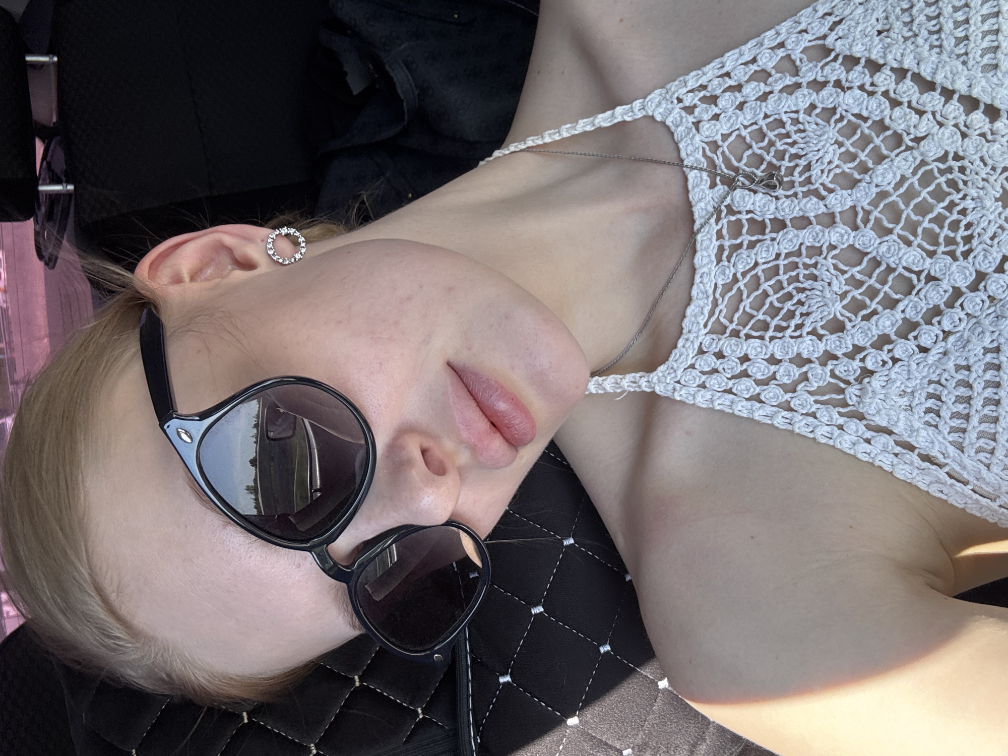 A light-skinned person with blonde hair is lying on a black quilted surface, possibly a car seat, wearing black sunglasses, a white crocheted top, and a silver necklace. The scene is lit by sunlight. The person is wearing a silver earring in their left ear. The reflection in the sunglasses shows what appears to be foliage and a vehicle, suggesting an outdoor or roadside setting. No further details about the location within the city of Perm, Russia, are discernible from the image.