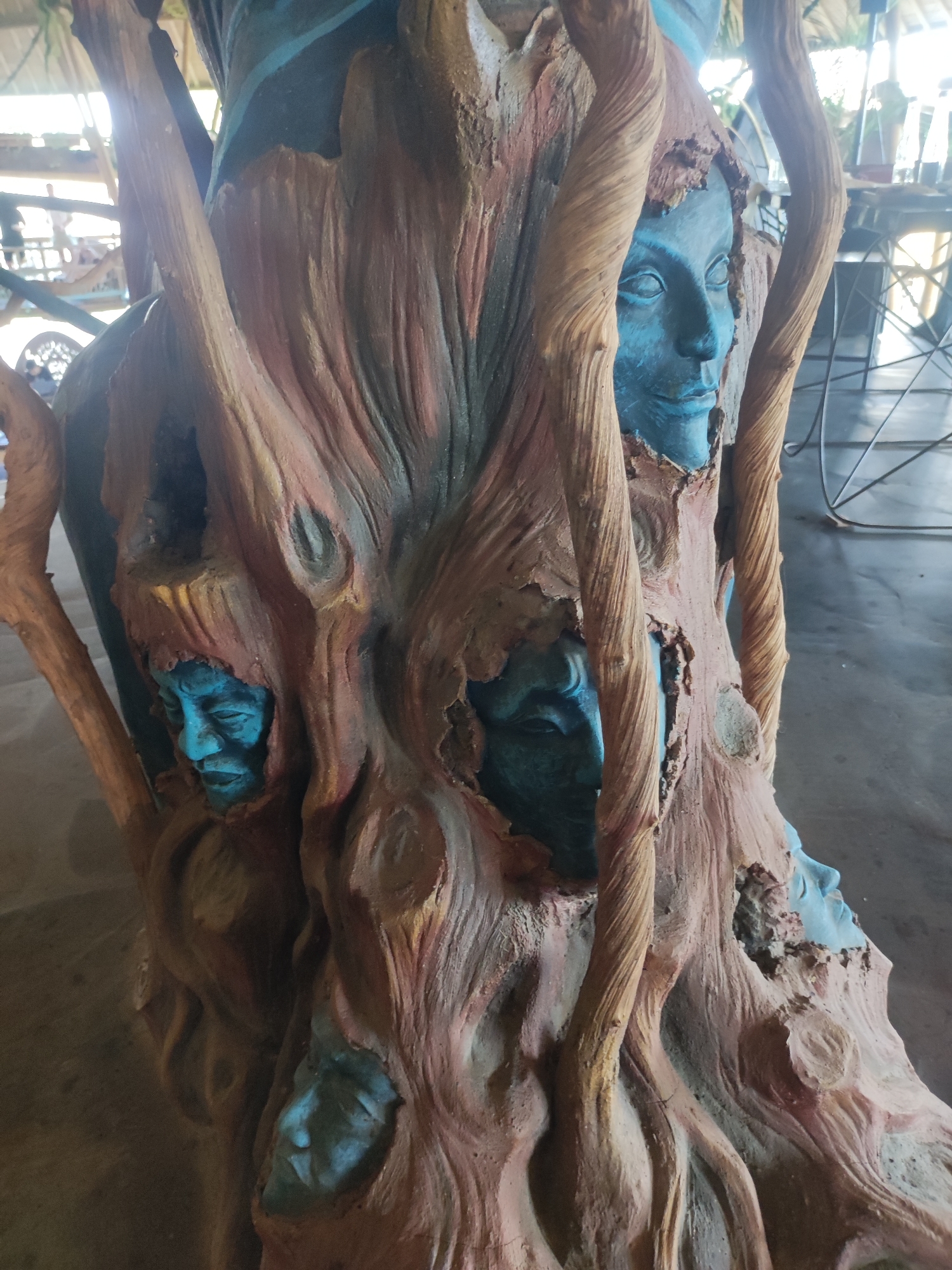 A medium-shot captures a crafted tree stump. Its external texture is intricately designed to resemble the ruggedness of tree bark. Imbedded within the bark are sculpted faces, painted in a vivid blue hue. The faces are positioned at different points and angles around the stump, each appearing embedded within the tree's exterior. The surrounding environment hints at an indoor space, with hints of tables and chairs. The image was taken in the city of Unknown, Indonesia.