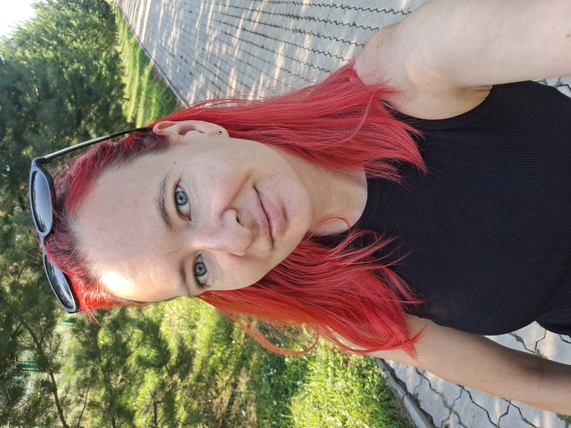 A medium shot shows a woman with bright red hair, wearing a black tank top and sunglasses pushed up on her head. She is outdoors on a paved walkway lined with trees. The woman is looking at the camera and smiling slightly. The background is a blur of green trees and a paved path. The image appears to be taken on a sunny day. This scene is located in the city of Atamanovka, Russia.